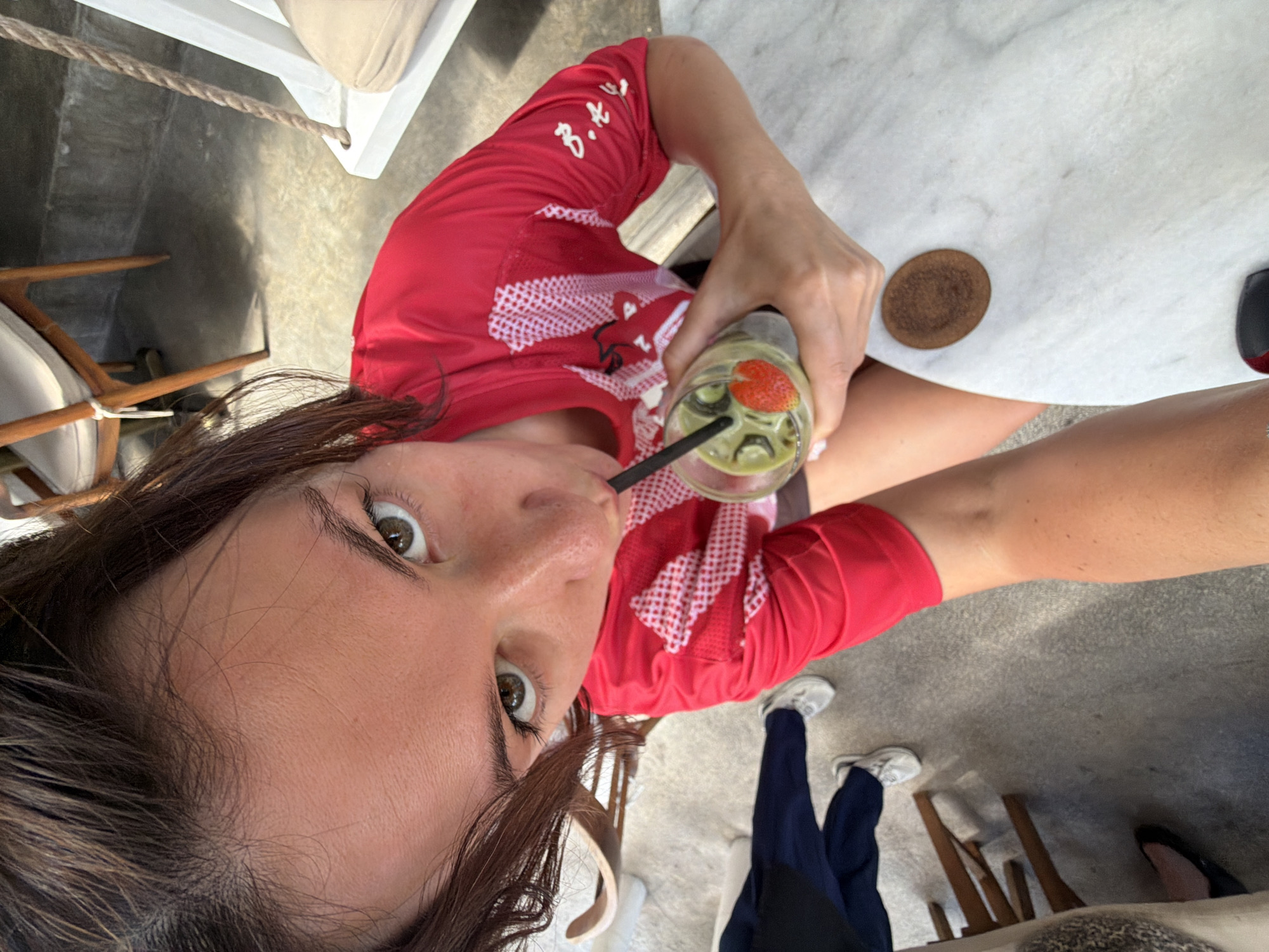 The image shows a high-angle, fisheye perspective of a person in the city of North Kuta, Indonesia. The subject, a woman with dark hair and green eyes, is wearing a red t-shirt with white detailing and is holding a glass of a green beverage with ice, a strawberry slice, and a kiwi slice. She is sipping the drink through a black straw. She is seated at a white marble table, on which a coaster is visible. In the background, a white swing chair with a tan cushion is partially visible, along with wooden chairs and a concrete floor. The overall environment suggests a casual outdoor cafe or seating area.