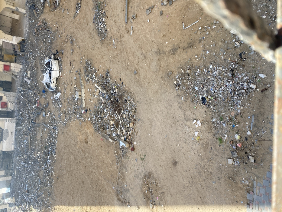 The image displays an extensive scene of urban devastation in Khan Yunis, Palestinian Territory, viewed from an elevated perspective. The foreground and midground consist of an unpaved, irregular expanse of compacted beige dirt, heavily strewn with scattered debris. This debris includes numerous small rocks, concrete fragments, twisted metal pieces, and various types of refuse.
In the upper-right quadrant, a severely damaged white four-door sedan is visible. The vehicle's roof is caved inward, windows are missing, and its body shows significant crumpling and dark scorch marks, particularly on the hood and passenger side. Near the center-left, a large pile of rubble contains multiple lengths of white, coiled plastic piping.
Along the top edge of the frame, a row of multi-story buildings stands in a state of severe disrepair. These structures exhibit extensive damage, including collapsed sections, numerous holes consistent with projectile impacts on their walls, missing windows, and exposed internal structural elements. Remaining visible architectural features show colors such as light grey, beige, red (from what appear to be window frames), and dark brown from scorching.
A small section of a paved walkway, constructed with interlocking pavers, is visible in the bottom-left corner. The bottom-right foreground is partially obscured by a blurred, rusted metallic object, likely a railing, indicating the photograph was taken from above the scene. No individuals are present in the image.