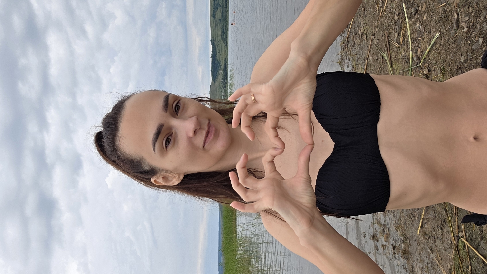 A young woman stands on the shore of a body of water, forming a heart shape with her hands in front of her chest. She is wearing a black bikini top. The background shows a wide expanse of water with a grassy shoreline and rolling hills in the distance. The sky is overcast with scattered clouds. The location is the village of Kupoyarovo, Russia.