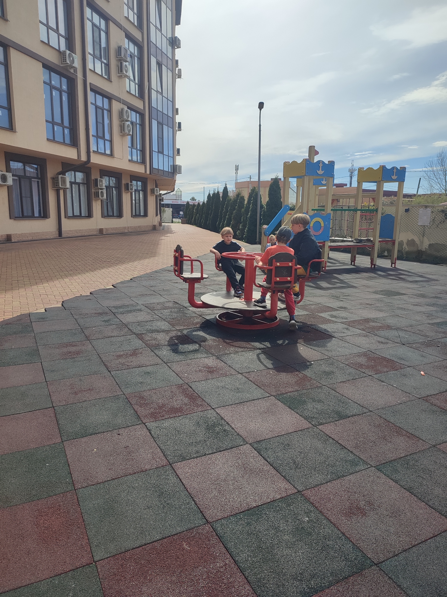 The image depicts a playground scene in Sochi, Russia. Three boys are on a red merry-go-round. One boy is wearing a black shirt and black pants. Another boy wears an orange shirt and red pants. The third boy is wearing a black jacket. The playground has safety flooring consisting of alternating red and dark grey square tiles. A playground structure with slides and ladders is visible in the background. Tall evergreen trees line a fence behind the playground. The building is beige with brown window frames and multiple air conditioning units. The sky is partly cloudy.