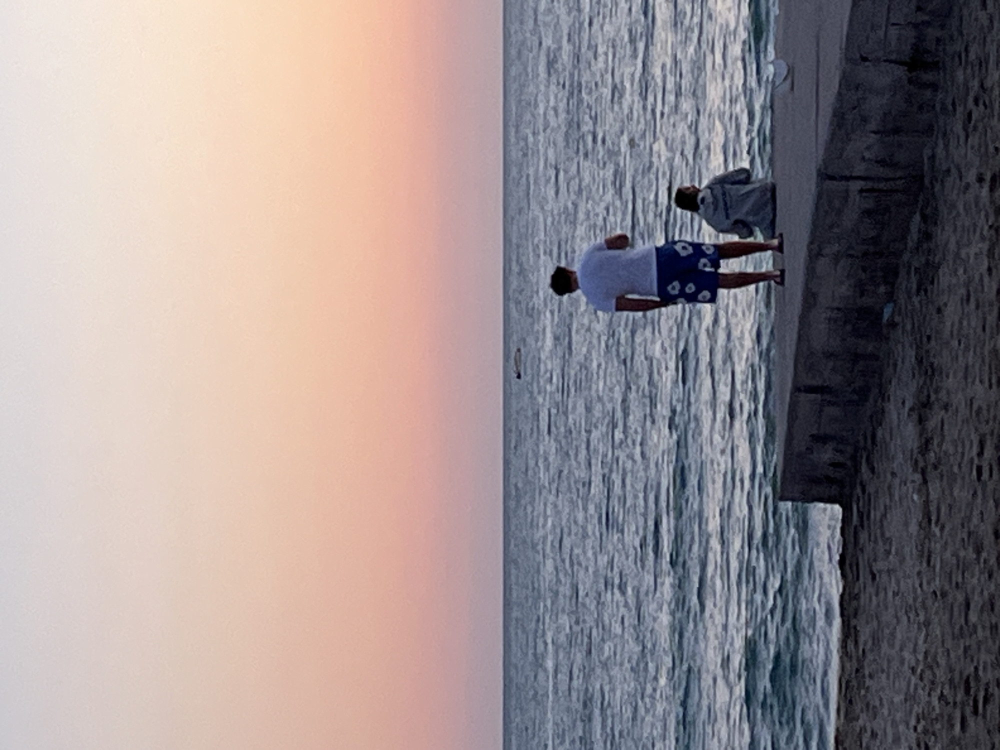 The image depicts a coastal scene, likely at dusk or dawn given the gradient of colors in the sky from white to orange.
The ocean is on the right side of the image, with waves. Two people are standing on a paved area adjacent to the water. One individual is wearing a white shirt and blue shorts. Another person is close by wearing a light-colored jacket.
The edge of the pavement meets a rough, dark, stone or concrete structure. The image suggests this could be a part of a breakwater or a retaining wall along the coastline.
Based on the prompt, this photograph was likely taken in Alanya, Turkey.