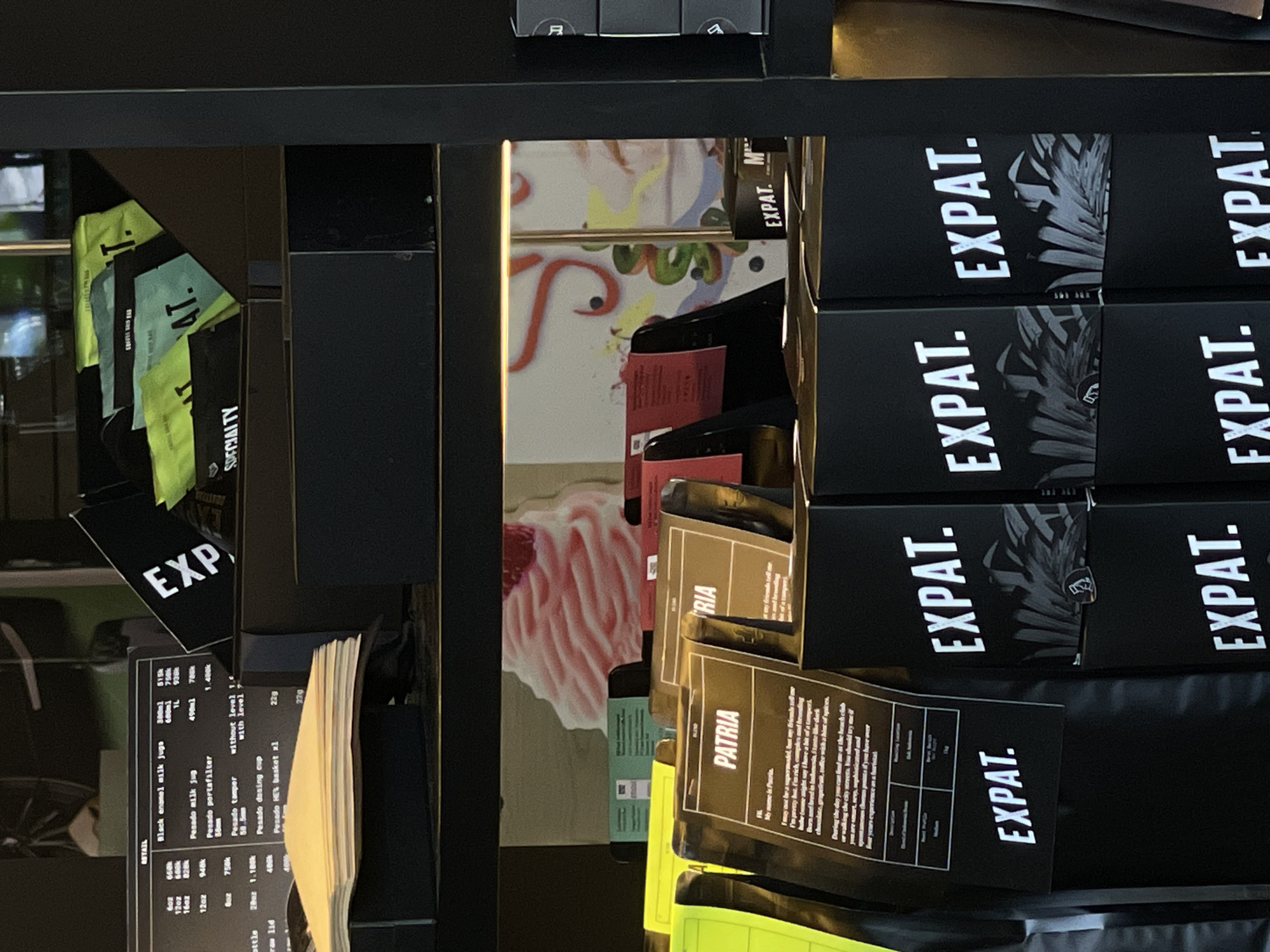 This image captures a well-organized display of coffee products and accessories, likely in a coffee shop or retail setting, in Kuta, Indonesia. The scene is dominated by dark shelves and black packaging, contrasted with vibrant green accents and a colorful background mural.
The main subjects are numerous packages of coffee, primarily branded "EXPAT." These black packages feature white, sans-serif text and often include a monochrome palm leaf graphic, suggesting a tropical or exotic origin. Several green packages, also bearing "EXPAT." or "AT." and "SPECIALTY" text, are visible on the left. Another prominent product line is "PATRIA," also in black packaging, which includes a detailed description. The "PATRIA" text describes the coffee as "rich, complex and rounding," "Born and bred in Indonesia," and mentions finding it at "the beach club or walking the city streets," aligning with the Kuta, Indonesia context.
Below the coffee bags, a printed menu or price list details various coffee accessories under the "RETAIL" heading. These include "Black enamel milk jugs" (300ml, 600ml, 1L priced at 515k, 930k), "Pesado milk jug" (12oz, 16oz at 490k, 786k), a "Pesado portafilter" (948k), "Pesado tamper" (58mm, with/without level at 750k, 1.180k), a "Pesado dosing cup" (229), and a "Pesado HE's basket xl" (274). A "Bottle" with a "raw lid" is also listed.
In the background, behind the displayed products, a vibrant, abstract mural or painting is visible, featuring large red, pink, yellow, and green brushstrokes and shapes, including a distinctive pink, swirled element that resembles a dessert or cupcake. No people or specific activities are shown, but the arrangement indicates a retail display setup, inviting customers to browse and purchase. The overall aesthetic is modern and sophisticated, with clear branding and detailed product information.