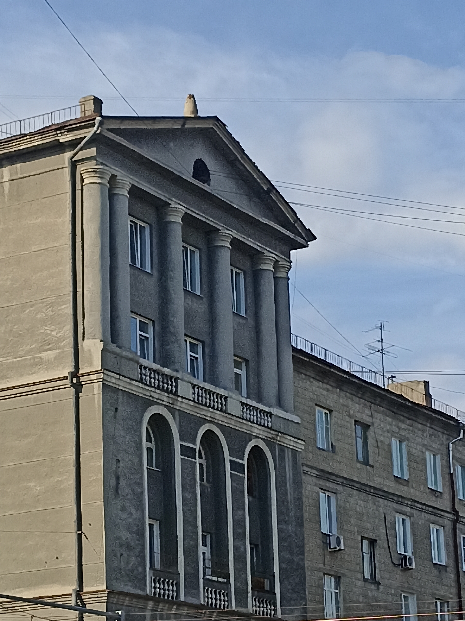 An exterior, eye-level, medium shot captures the facade of a multi-story building in Novosibirsk, Russia. The building, constructed of gray material with a textured finish, features prominent classical architectural elements. The left side of the building displays a large, grey structure with a triangular pediment adorned with decorative columns and a decorative railing. Below the pediment, the building has several windows with white frames. The right side of the building is slightly lower and appears to be a separate structure attached to the first. This section is characterized by a facade of smaller, block-like stones and has numerous windows with white frames and several air conditioning units mounted on the exterior walls. The sky above is a mix of blue and white clouds, with several overhead power lines crossing the scene. The overall impression is one of a Soviet-era residential or administrative building.