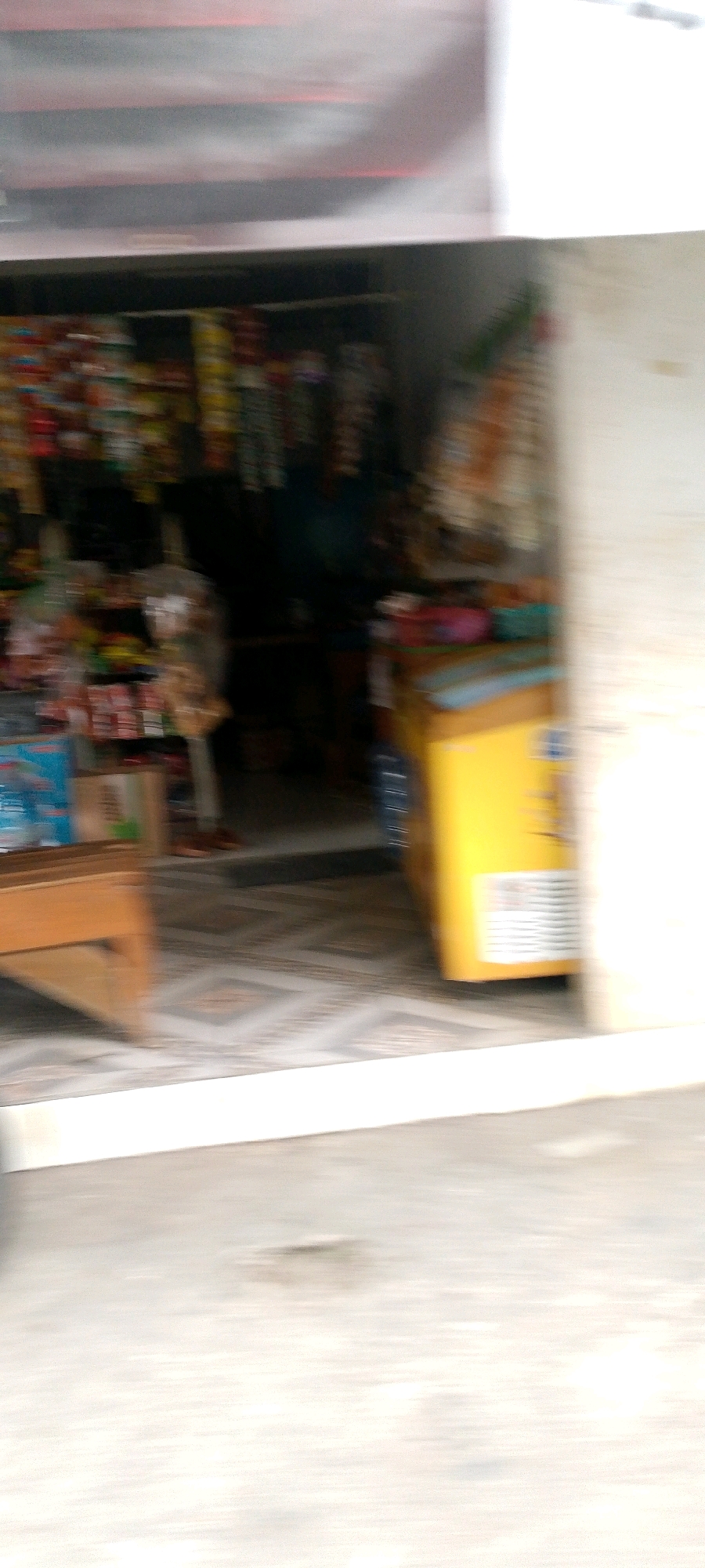 The image captures the entrance of a small store or warung, likely in Langensari, Indonesia. The scene is characterized by motion blur, suggesting the photo was taken from a moving vehicle. Inside the store, shelves are stocked with various packaged goods, appearing to be snacks and drinks. A prominent yellow refrigerator or freezer unit is visible on the right side, also with some products on top. A wooden counter or display is partially visible on the left, with some items on it. The floor inside the store has a patterned tile design. The outdoor area in front of the store shows a paved surface, and the overall environment suggests a commercial setting within a town or city. There are no people clearly visible due to the blur.