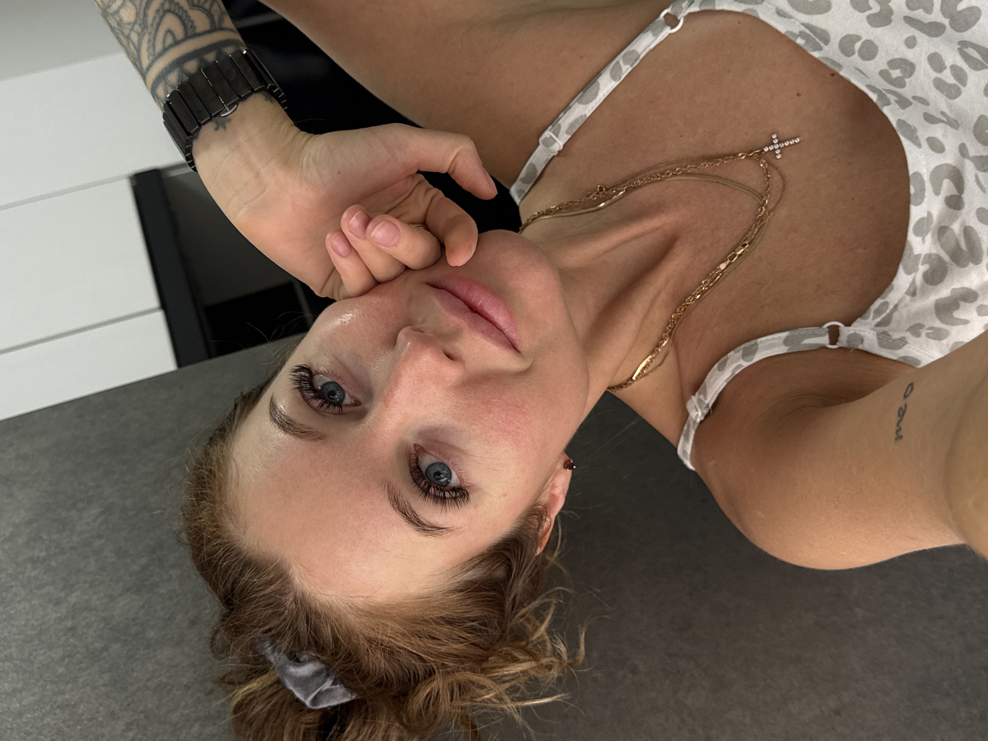 An overhead, slightly rotated shot captures a woman lying on her back, looking directly into the camera. She has blue eyes, long eyelashes, and light brown, curly hair pulled back, with some strands framing her face. Her expression is neutral and contemplative. She wears a light-colored top with a grey and white animal print, possibly a camisole or tank top.
Her left hand rests near her chin, with her fingers gently curled. On her left wrist, a dark, segmented wristwatch is visible, along with an intricate mandala-style tattoo on her forearm. A subtle, small star-like tattoo can also be seen beneath the watch. A gold-toned, multi-layered necklace with a small, sparkling cross pendant adorns her décolletage. On her right inner forearm, a small script tattoo reads "meo." Her skin appears dewy and well-hydrated, with what looks like a natural lip color or lip blush.
She is lying on a plain, dark grey surface, possibly a bed or couch, with a white paneled wall visible in the background, suggesting an indoor setting in Chaikovsky, Russia. The lighting is soft and even. A grey scrunchie or hair tie is visible in her hair on the left side of her head.