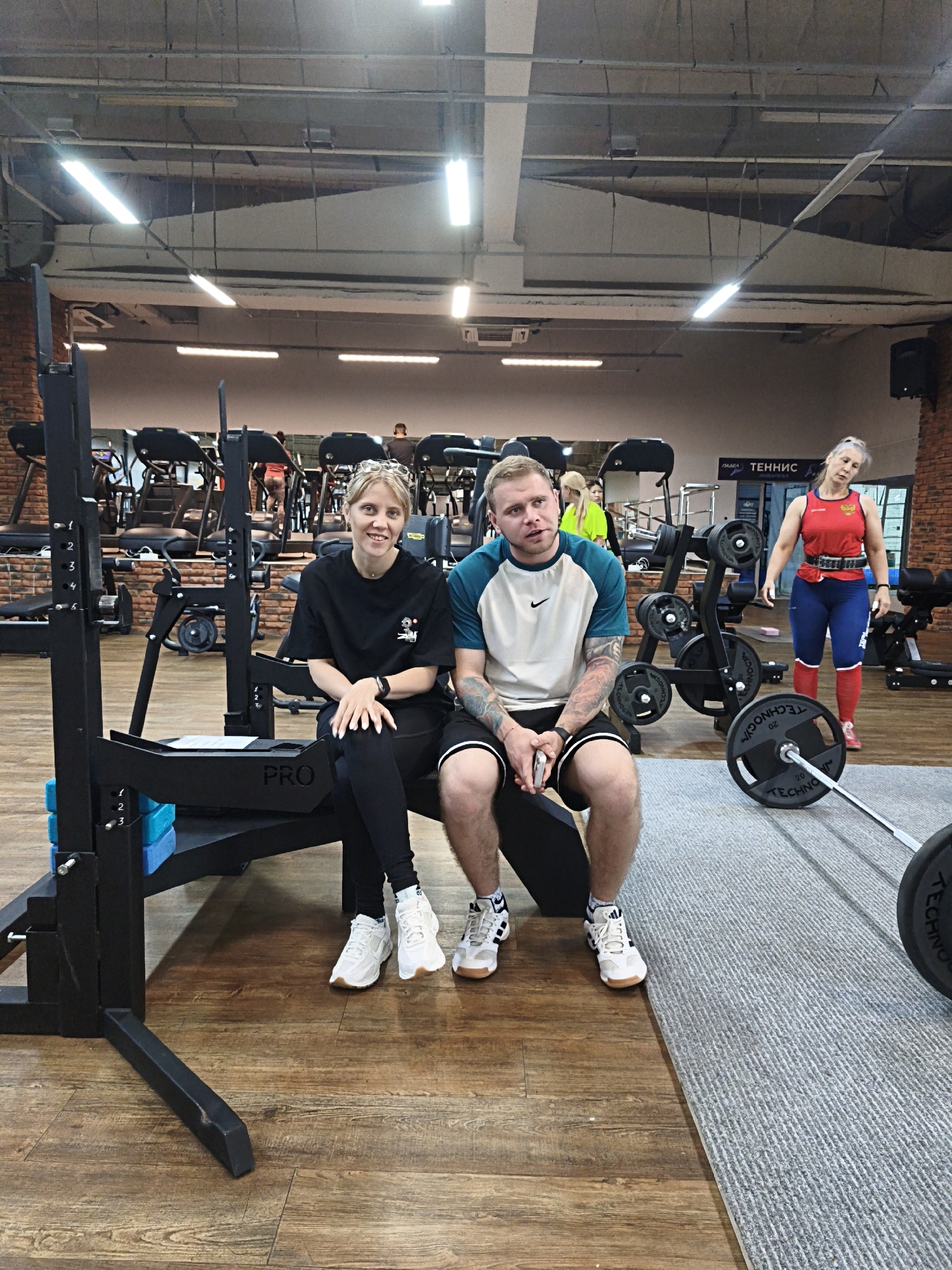 The image depicts an indoor scene in a gymnasium, presumably in the city of Chaikovsky, Russia, as indicated by the location.
Two individuals, a woman and a man, are seated on a piece of weightlifting equipment labeled "PRO." The woman, on the left, has fair hair, wears a black t-shirt, black leggings, and white sneakers. The man beside her has short blond hair, a light-colored t-shirt with dark green accents and a Nike logo, dark shorts, and sneakers with a predominantly white color scheme. He has visible tattoos on his left arm.
In the background, a woman in red and blue sportswear stands near weightlifting equipment with plates and a barbell. Other gym equipment, including treadmills and weight machines, are arranged behind the seated individuals. The gym's interior features brick walls, a wooden floor (except where a grey mat is), overhead lighting, and exposed ceiling beams.