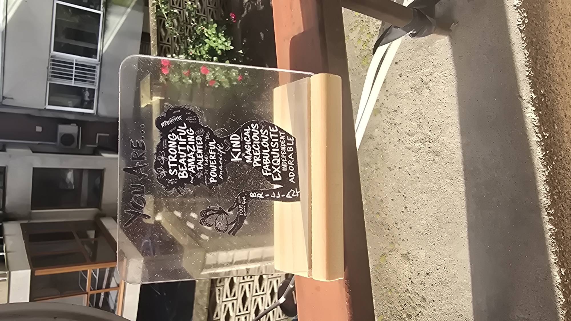 An outdoor, well-lit photograph features a transparent acrylic plaque bearing a motivational message, supported by a light-colored wooden block. The plaque, positioned on what appears to be a balcony or outdoor ledge, prominently displays the phrase "YOU ARE..." on its left side. The central design is a black silhouette of a woman's head and upper torso, filled with uplifting white text. These adjectives include "STRONG, BEAUTIFUL, AMAZING, TALENTED, KIND, POWERFUL" on the left side of the silhouette, and "KIND, MAGICAL, PRECIOUS, FABULOUS, EXQUISITE, INDEPENDENT, ADORABLE" on the right. A small butterfly graphic is visible below the silhouette, accompanied by the word "YOURS" and the initials "BR-LJ-AY".
The background reveals an urban setting, likely in Varna, Bulgaria, with a multi-story building featuring several windows, some with air conditioning units, visible on the left. To the right and behind the plaque, lush green foliage with vibrant red flowers adds a natural element. The ground beneath appears to be rough concrete, partially shaded by the wooden support, indicating bright daytime conditions with direct sunlight. The overall impression is of a personalized, inspirational decorative item displayed against a typical city backdrop.