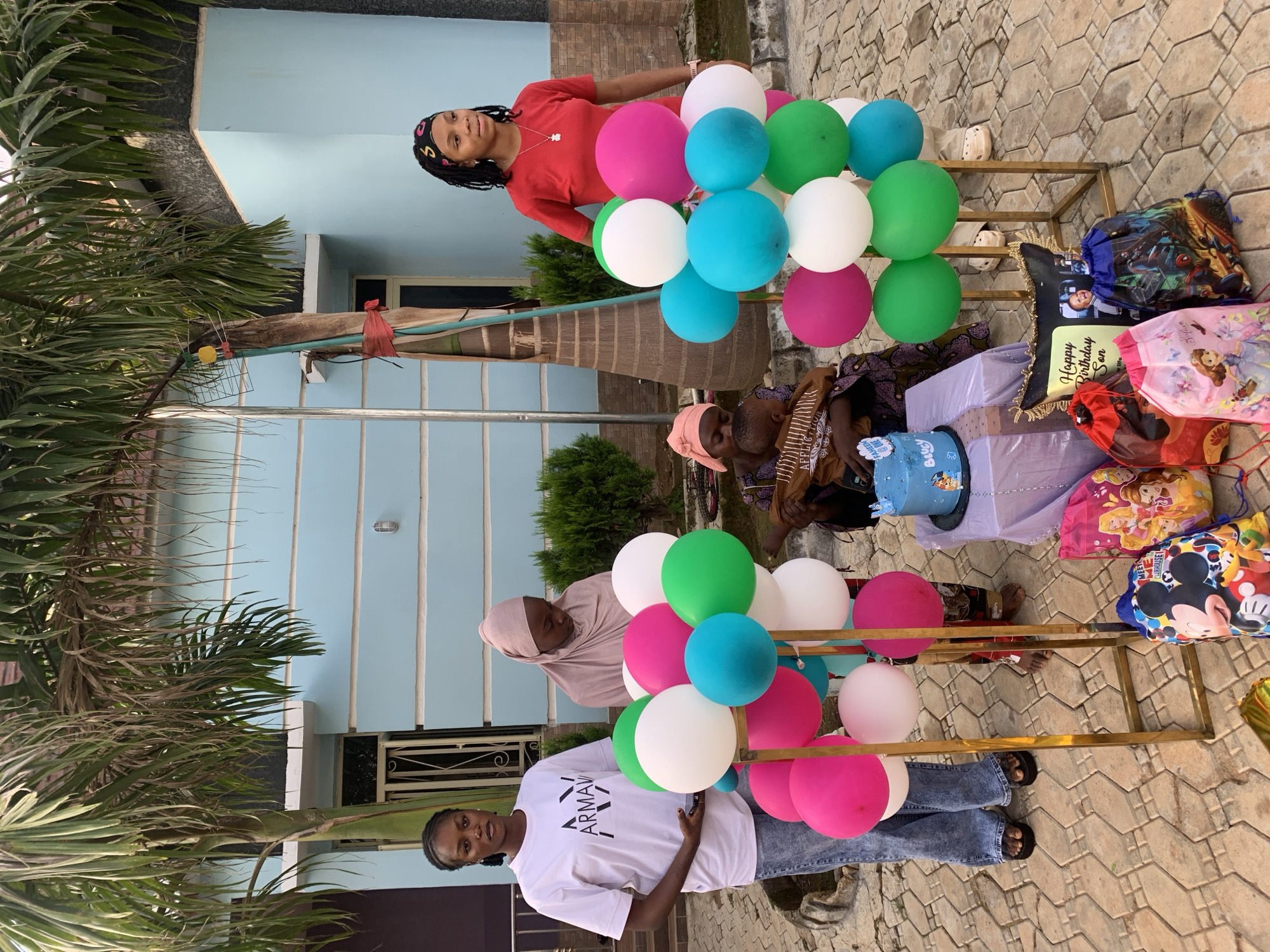This image depicts an outdoor celebration in Jos, Nigeria, featuring five individuals and birthday decorations positioned in front of a light blue residential building. The ground is paved with interlocking reddish-brown concrete pavers.
Five people are present: three adult women, one woman holding an infant, and the infant.
1. On the far left, an adult woman with dark braided hair stands, wearing a white t-shirt with an "ARMANI" logo, light blue jeans, and black flip-flops. She holds a dark object, likely a mobile phone, in her left hand and looks towards the camera.
2. Behind the left balloon stand, an adult woman in a light pink hijab and matching traditional attire stands, looking downwards.
3. In the center-left, an adult woman wearing a patterned dress and a pink headscarf is seated on a low stool. She holds an infant, wrapped in a brown and white patterned cloth, whose head is visible facing away from the camera.
4. On the far right, an adult woman with dark dreadlocks and a colorful headband stands, wearing a red t-shirt, light-colored pants, and white clogs. She wears a necklace with a small pendant and looks slightly to her right.
5. The infant held by the seated woman is partially visible, appearing to be an infant or toddler.
Two tall, gold-colored metal stands are positioned in the foreground, each displaying clusters of balloons in pink, white, green, and light blue. Between these stands, a small table draped in purple fabric holds a two-tier light blue birthday cake. The cake features a character resembling Bluey, the text "SAHEED ONE", and a small card stating "Baby". On the ground around the cake table are multiple colorful drawstring gift bags, depicting characters such as Mickey Mouse, Disney Princesses, Sofia the First, and Spider-Man. A decorative pillow with "Happy Birthday Son" and a photographic inlay is also on the ground.
The background features a light blue, two-story building with white window frames and balconies. Large palm trees frame the scene on both sides, with dense green foliage visible behind the balloon stands. A yellow object and red tape are visible attached to the trunk of the right palm tree. The natural light indicates a clear day.