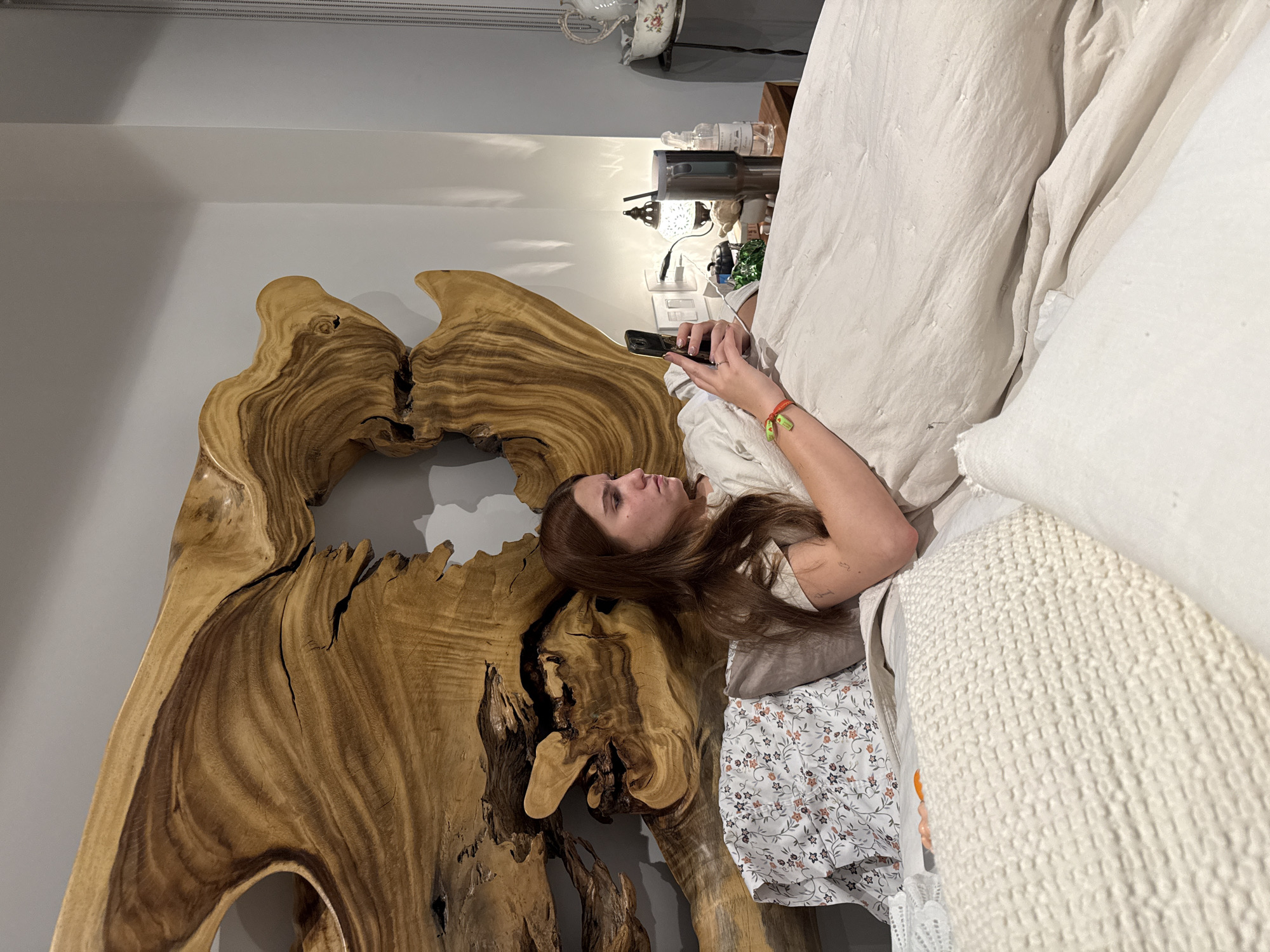 A woman lies in a bed, using a smartphone. She is positioned in a room with a unique, large wooden headboard. The location is Huixquilucan de Degollado, Mexico. The room also contains a lamp, bedside table, and other bedroom furnishings.