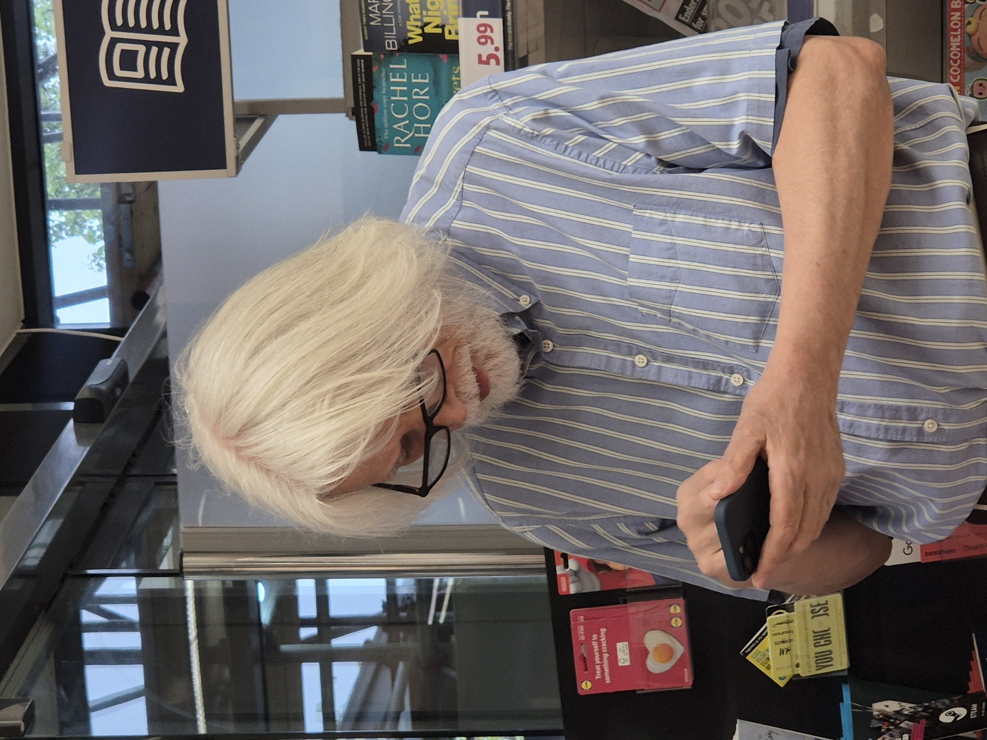 An older man with long white hair and a white beard wearing glasses and a striped blue shirt is looking down at his mobile phone while standing in a bookstore. Behind the man, there is a sign with a book graphic, some books, and other products.