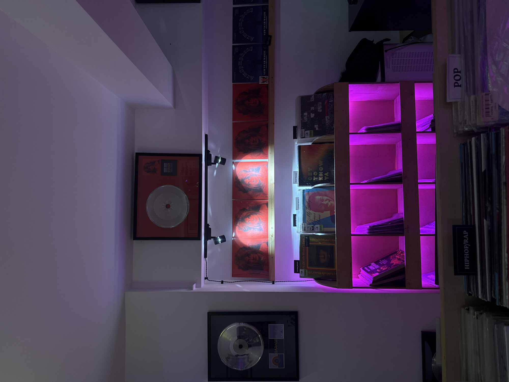 This indoor scene presents a vertically-oriented view of a shelf arrangement, likely within a record store or personal collection. The lighting is prominent, with a pink hue emanating from behind a wooden shelving unit to the right. On the left wall, framed records or memorabilia are displayed, illuminated by a small adjustable spotlight. The center shelf holds a series of records with discernible album art. Labels indicating genres such as "POP" and "HIPHOP/RAP" are attached to the rightmost shelf, suggesting a categorization system. The OCR content hints at album names and genres, and a label that says "Garage".