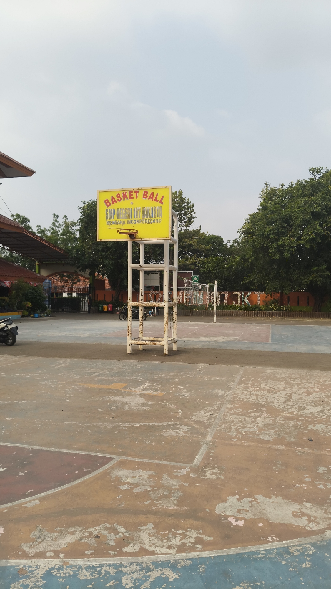 The image depicts an outdoor basketball court at SMP Negeri 107 Jakarta, located in South Jakarta, Indonesia. In the foreground, a weathered white basketball hoop stands on a court with faded red and blue markings. Behind the hoop, a yellow sign reads "BASKET BALL" in red letters, with "SMP NEGERI 107 JAKARTA" and "MEMBINA KORPU SESANO" below it. The court area includes a net and the inscription "JKT" painted on an orange wall in the background. To the left, a building with a brown roof and an arched entrance is visible, along with a parked white motorcycle. Trees line the perimeter of the court. The sky is overcast.