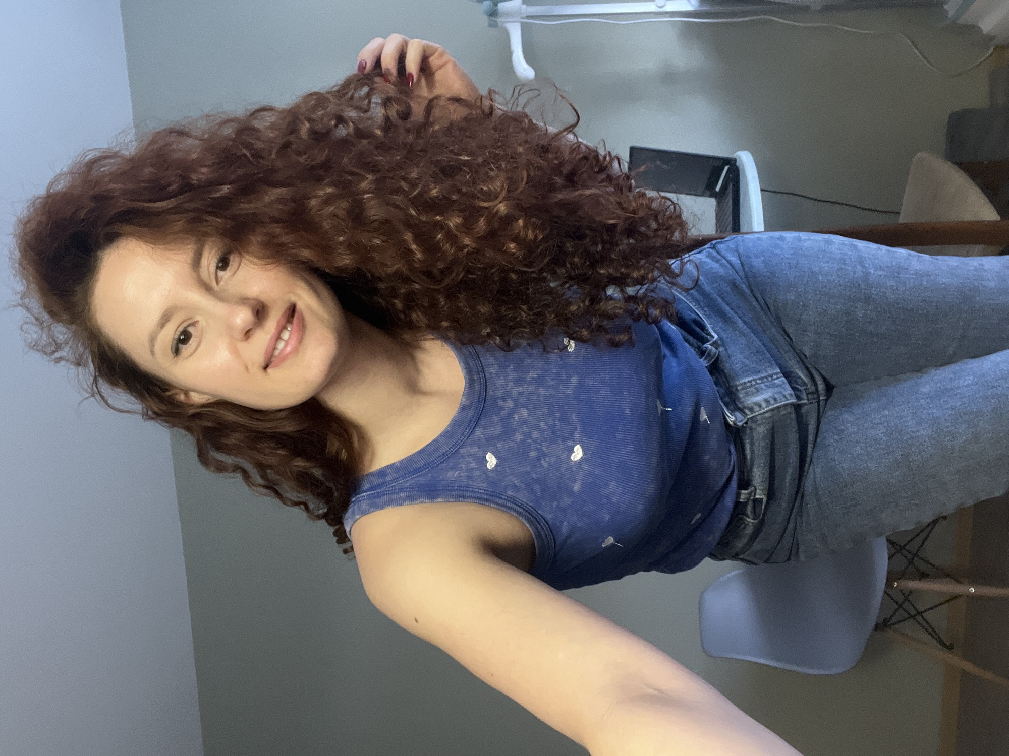 A woman with red curly hair, wearing a blue tank top with white hearts and light blue jeans, is sitting on a light blue chair. The photo appears to have been taken indoors, with a desk and some other furniture visible in the background. The location is not discernible from the image.