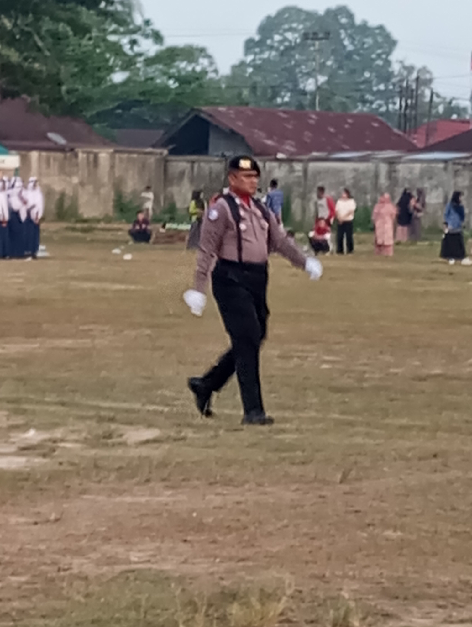 In Tanjung Gading, Indonesia, a man in a police uniform with a black beret, a light brown shirt, and black pants walks across a grassy field. He is wearing white gloves and black shoes. Behind him, several people are gathered, some appearing to be students in uniform, while others are dressed in civilian clothing. Buildings with reddish roofs and a concrete wall are visible in the background. The environment suggests an outdoor event or ceremony taking place.