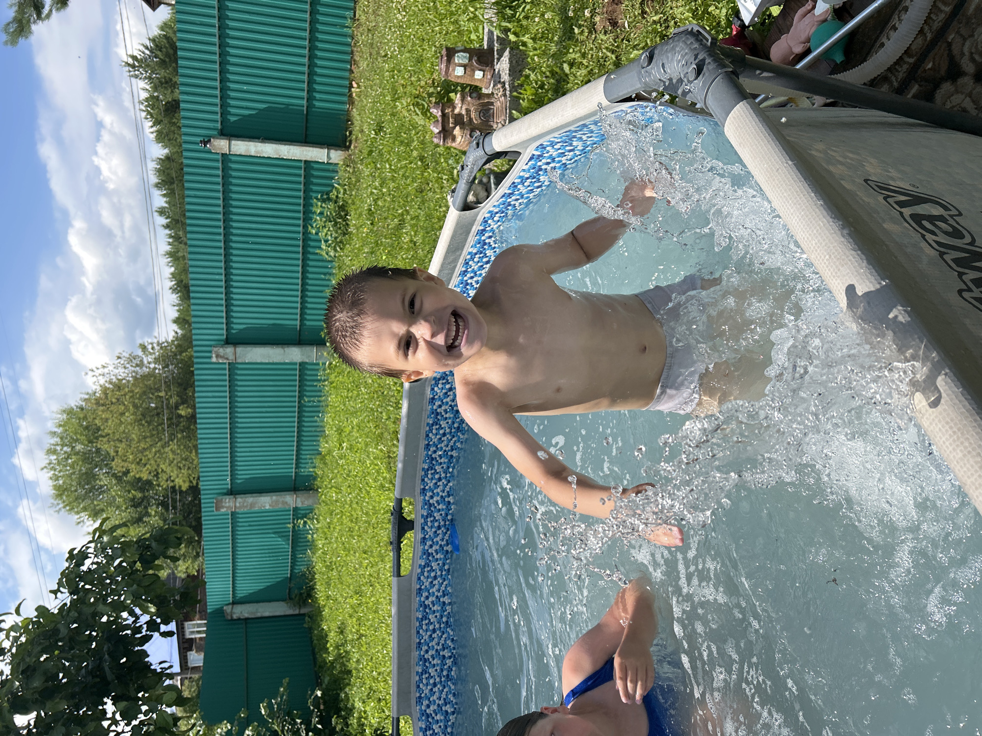 A young boy is splashing joyfully in a small above-ground swimming pool. He is shirtless and wearing light-colored swim trunks. Water sprays around him as he moves his arms. Another child, only their shoulders and head are visible, is in the pool with him, wearing a blue swimsuit. The pool has a blue and white patterned liner at the top edge and is filled with clear blue water.
The scene is set outdoors on a sunny day, with lush green grass and trees in the background. A green corrugated metal fence runs along the left side of the frame. The time of day appears to be daytime, likely afternoon, given the bright sunlight casting shadows. The boy's expression is one of pure happiness and excitement, with a wide grin and open mouth. The overall atmosphere is one of carefree summer fun and childhood enjoyment. The location is identified as Foki, Russia. There is visible text on the side of the pool, which reads "Bestway".