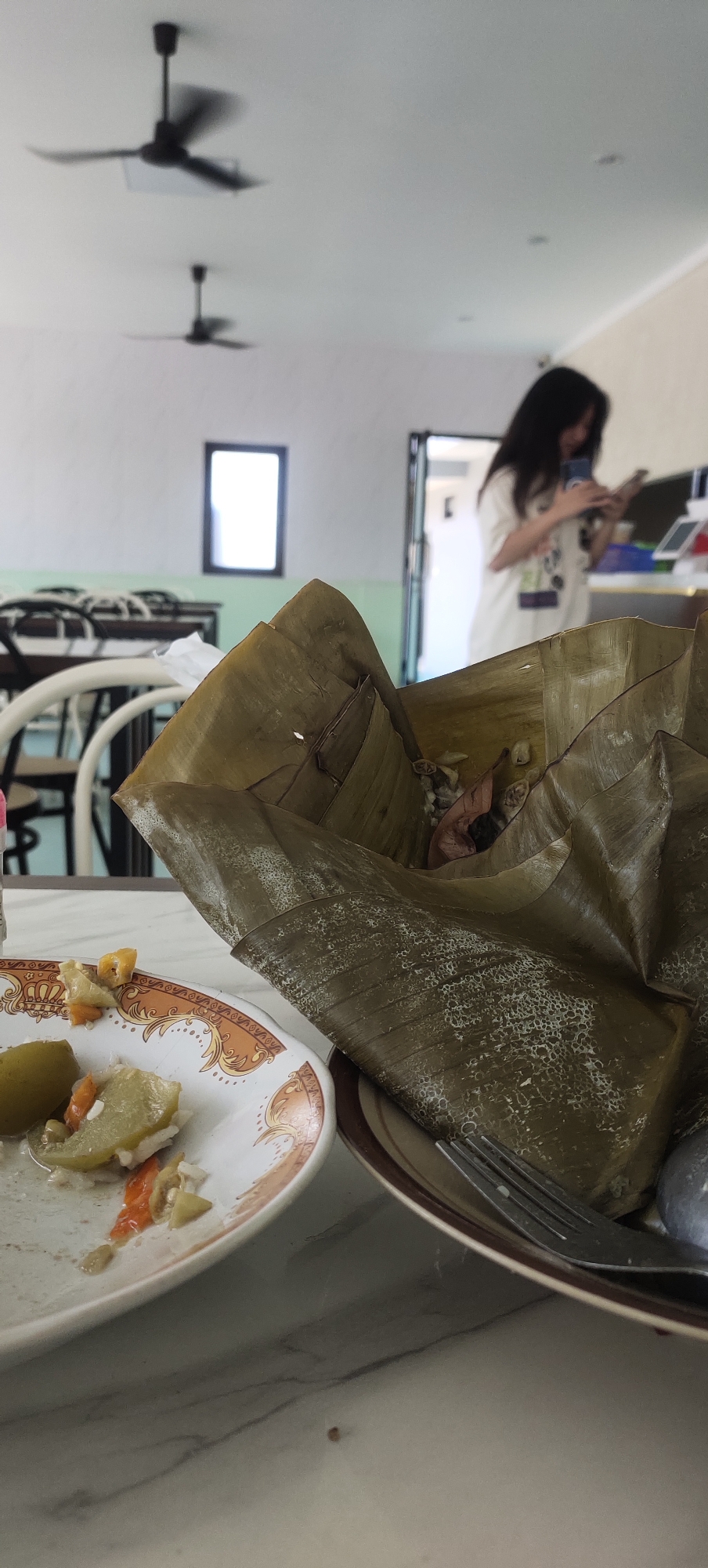In the city of Jatirejo, Indonesia, on a marble-patterned table, a partially eaten meal is presented. To the left, a white plate with an orange and gold rim holds remnants of food, including pieces of green vegetables, what appears to be orange pepper, and white rice. To the right, a large serving of food wrapped in banana leaves is the main subject. The leaves are a muted green, with a textured surface, and have been folded to enclose the contents. Peeking from the top of the banana leaf wrapping are glimpses of rice and some dark, indistinct ingredients. A fork rests on the plate to the right, next to the banana leaf-wrapped food. In the background, a woman with long dark hair, wearing a white t-shirt, is partially visible, looking down at her phone. She is standing near an open doorway. Above, two black ceiling fans are mounted on a white ceiling. The environment appears to be an indoor dining area, with the light suggesting daytime.