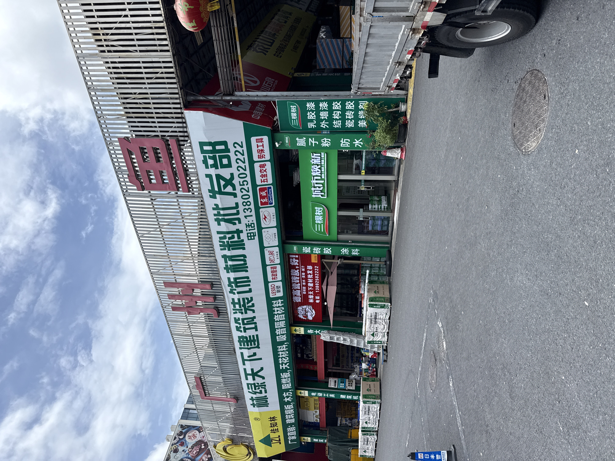 The image shows a street scene in Tianhe District, China, with a building that appears to be a wholesale store for building and decorative materials. The storefront has large signage with Chinese characters, some of which are in red. The store is offering various products like architectural molds, wood, fireproof panels, terrazzo, and sound-absorbing materials. There are several stacks of boxed materials and a truck parked in front of the building, suggesting delivery or loading activity. The street is paved with asphalt, and there are some markings and a manhole cover on the ground. The sky is cloudy.