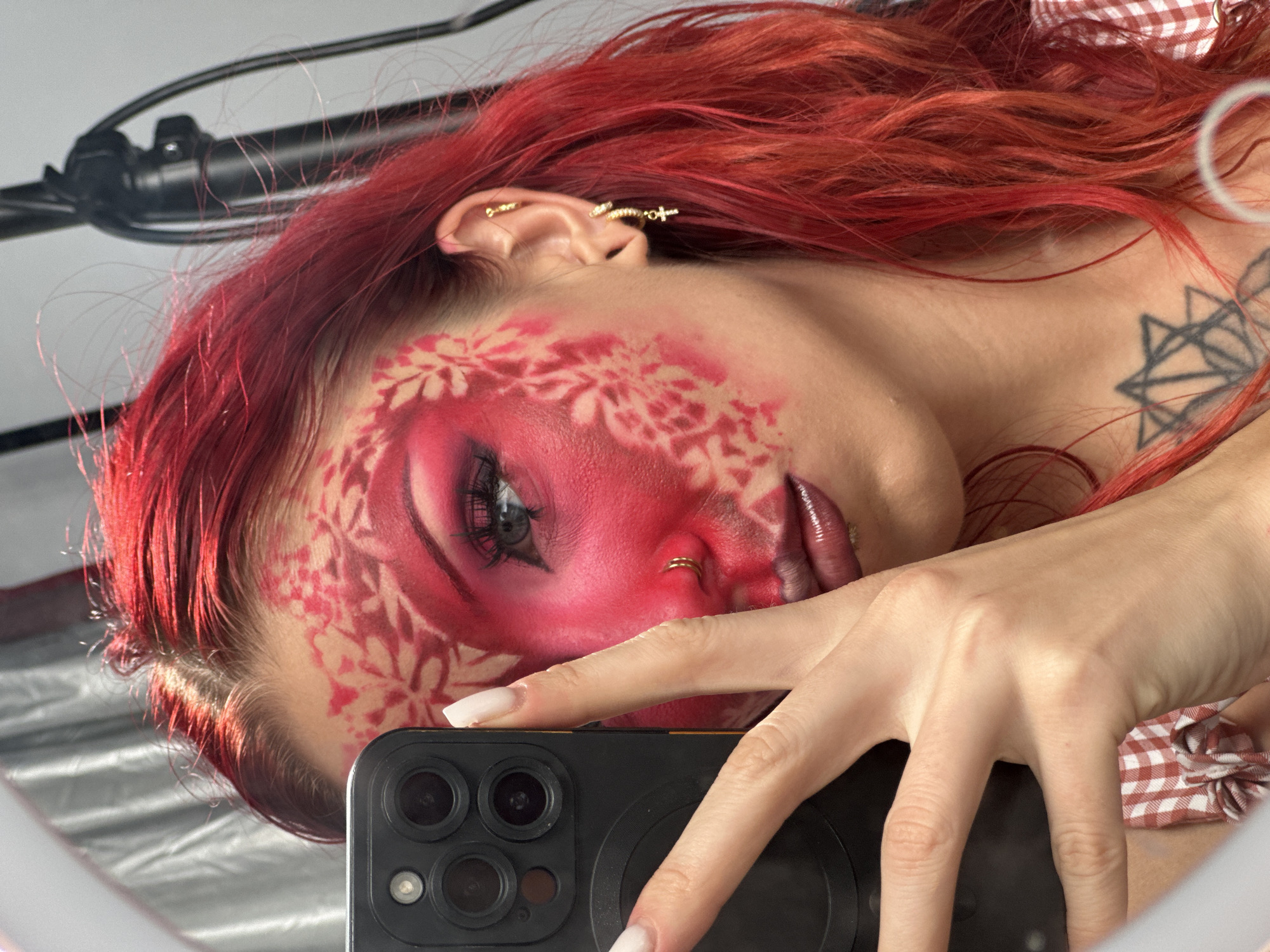 A young woman with vibrant red hair takes a selfie in a mirror. She is wearing a red and white checkered shirt. Her face is adorned with intricate red and pink makeup, with a floral pattern on her left cheek and temple. She has dramatic eye makeup, including pink eyeshadow and false eyelashes. She is also wearing a nose ring and multiple earrings, including one with a cross charm. She holds a black smartphone in her left hand, obscuring part of her face. The background appears to be a studio setting with a ring light visible at the top left. There is a tattoo on her chest, which is a geometric star shape. The image does not contain any information about the location in North Kuta, Indonesia.