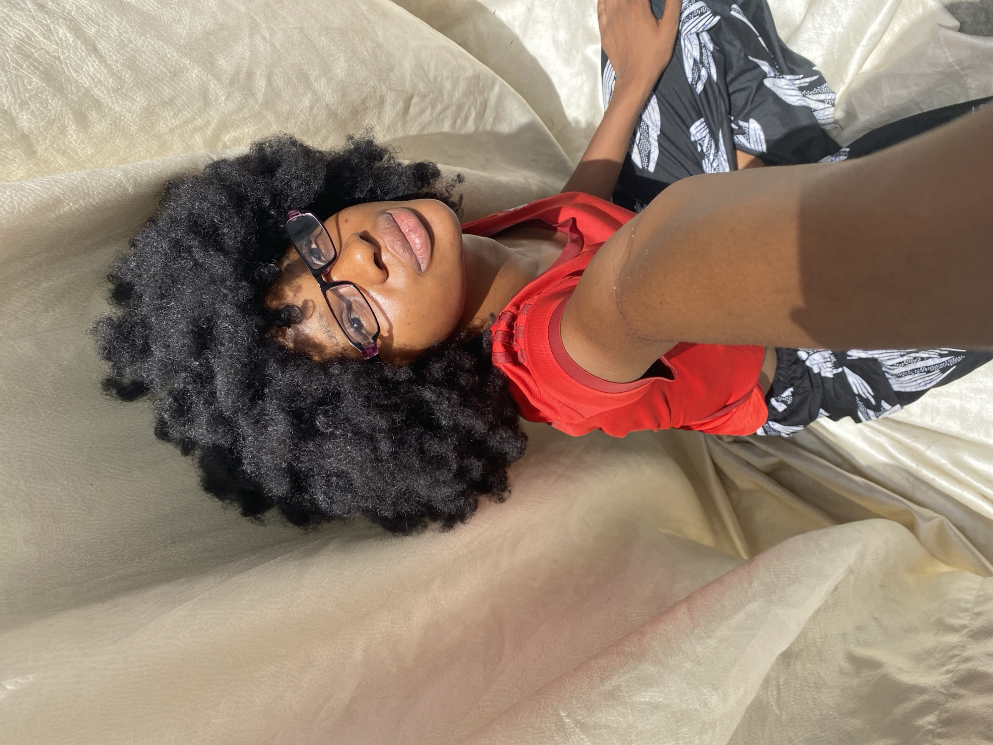 A Black woman with dark, curly hair and glasses lies on a tan fabric surface. She wears a red shirt and patterned pants. Her arm is extended toward the camera. The location is not specified.