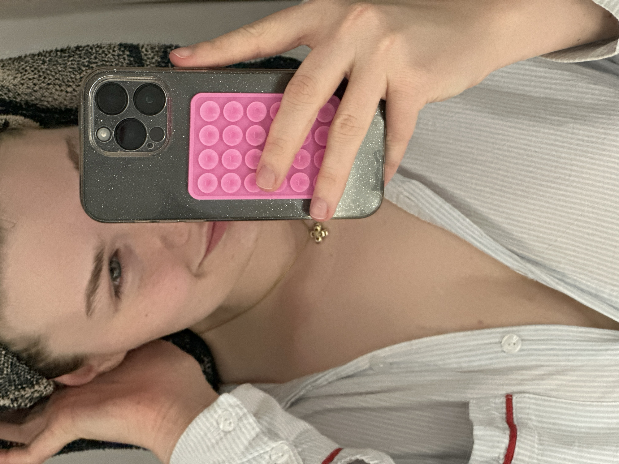 A person is taking a selfie while lying down. The person is holding a smartphone with a sparkly grey case and a pink pop-it accessory. The camera lens of the phone is prominently visible. The person has fair skin, light hair, and is wearing a light-colored striped shirt with red trim. A gold necklace with a clover-shaped pendant is visible on their neck. The background consists of a patterned fabric and part of the person's shirt. The location of the photo is not discernible within the image. There is no evidence to suggest it was taken in Kazan, Russia.