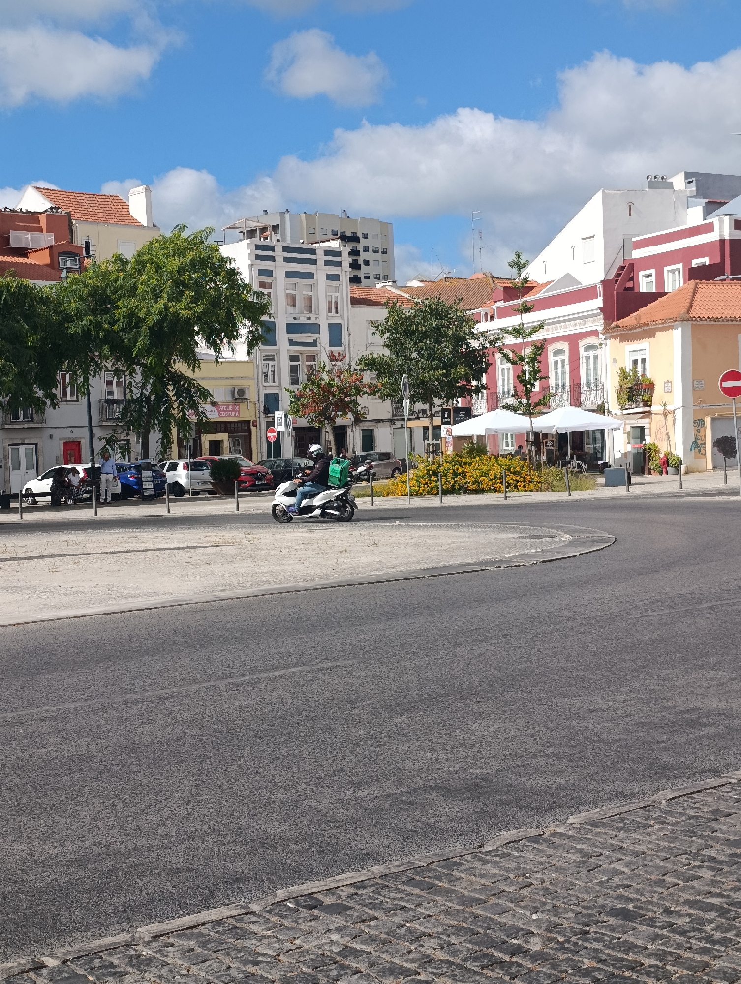 In Setúbal, Portugal, a street scene unfolds under a bright, cloudy sky. The foreground is dominated by a paved road and a cobblestone sidewalk. A white scooter with a green delivery bag is being ridden by a person wearing a helmet. In the middle ground, several cars are parked along the street, and a row of buildings with colorful facades, including red, yellow, and white, line the background. Trees with green foliage are visible, and two white umbrellas are set up in front of a building, suggesting an outdoor seating area. The architecture is typical of a European city, with multiple stories and balconies. A red and white circular sign with a red horizontal bar is affixed to a pole on the right side of the image.