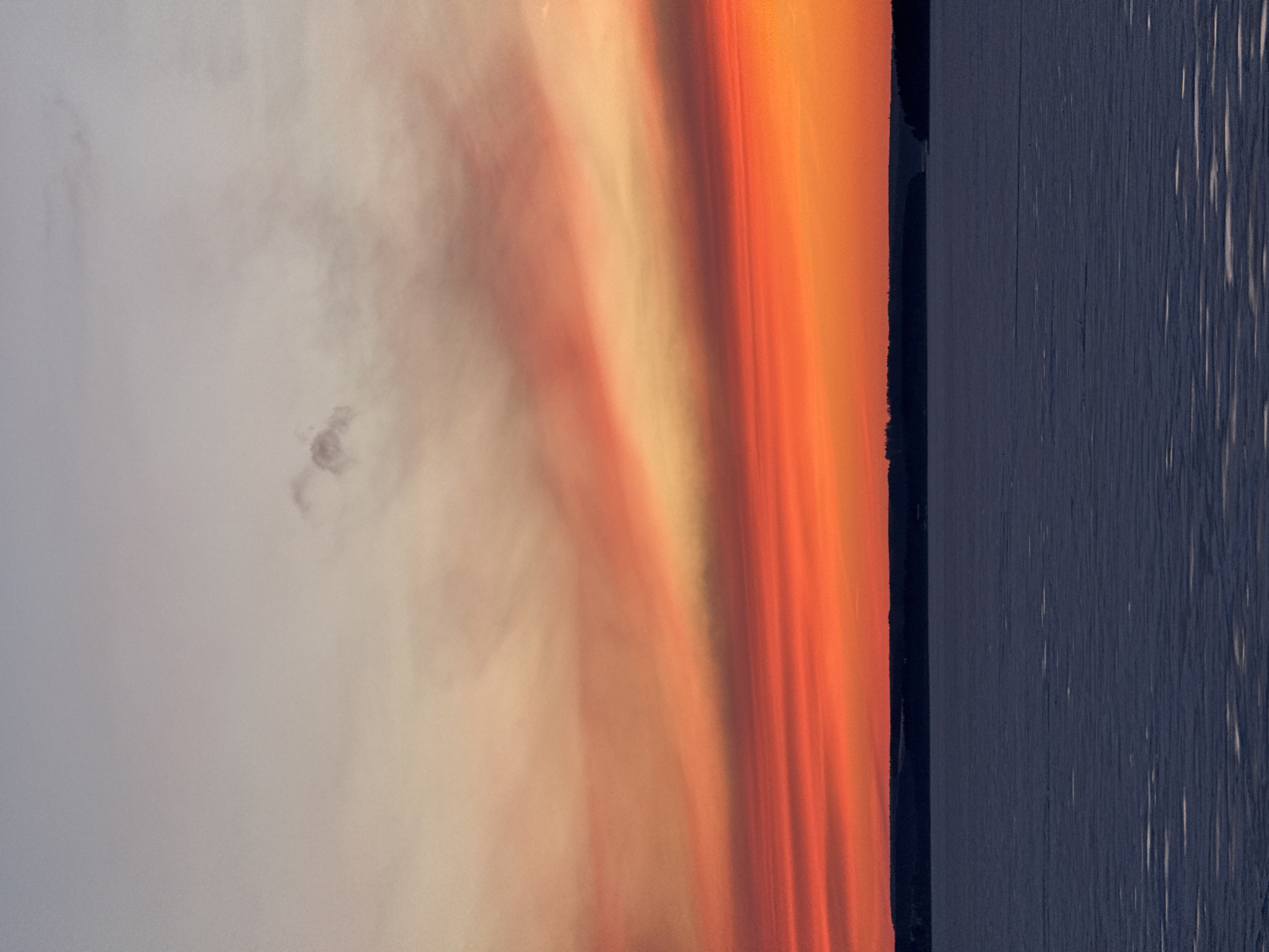 The image depicts a landscape at dusk, likely near a body of water. The sky transitions from light grey with cloud formations to vibrant shades of orange and red. A dark, vertical band of either land or a structure sits at the right edge of the frame, separating the sky from what appears to be a body of water with vertical, possibly reflective, streaks. The overall impression is of a calm, scenic view during twilight. While this appears to be a sunset over a body of water, there is nothing in the photo to conclusively link it to the specific location of Votkinsk, Russia.