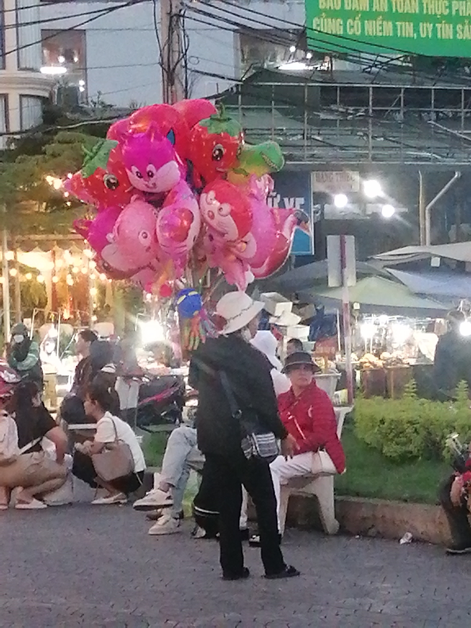 Captured in Da Lat, Vietnam, this eye-level shot showcases a bustling street scene at night. The foreground is dominated by a bouquet of vividly colored, animal-shaped balloons, primarily pink and red, hinting at a vendor selling them.
A diverse group of people populates the scene. Several are seated on the ground, while others are seated on stone benches, suggesting a casual gathering or market setting. One individual, donning a white hat, is dressed in black pants and a black jacket. A woman in a red jacket is seated on a bench.
In the background, vendor stalls are illuminated, indicating the presence of a lively marketplace. Above, a green sign with text, partially obscured, is visible. Electrical lines crisscross overhead, adding to the urban feel of the location. The building in the background is multi-story with visible windows and lighting. The stone or concrete walkway in the foreground contrasts with the lush green plants.