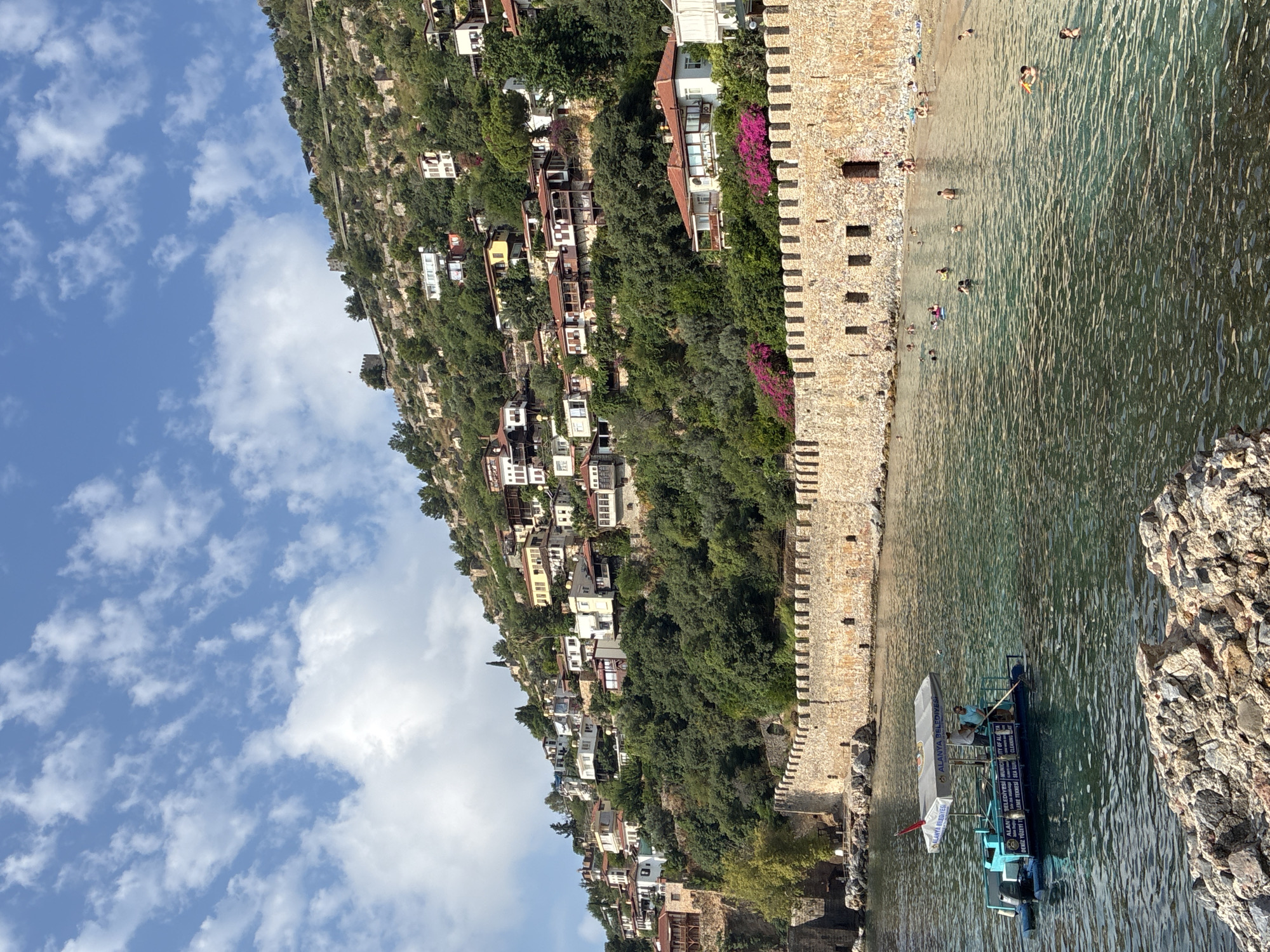 The image depicts a coastal scene in Alanya, Turkey, featuring a historical stone wall bordering a body of water. The wall, likely a remnant of the city's ancient fortifications, has an crenellated top and appears to be constructed from large, rough-hewn stones. Beyond the wall, a hillside is densely populated with numerous houses, varying in architectural style and color, cascading down towards the sea. The houses are interspersed with lush green trees, suggesting a Mediterranean landscape.
In the foreground, the water is clear and turquoise, revealing the rocky seabed in some areas. Several people are visible swimming and relaxing in the water and on a small sandy beach adjacent to the wall. To the left of the image, a small boat with a canopy is present. The boat appears to be a cleaning vessel, as evidenced by the signage on its side which includes text like "ALANYA BELEDIYESI" (Municipality of Alanya) and "SEA CLEANING." Two individuals are on the boat, one of whom is operating a pole, likely to collect debris from the water.
The sky above is a bright blue with scattered white clouds, indicating fair weather. The overall impression is one of a scenic and historically significant location being enjoyed by locals and possibly tourists. The combination of ancient architecture, natural beauty, and modern activity creates a dynamic and engaging scene.