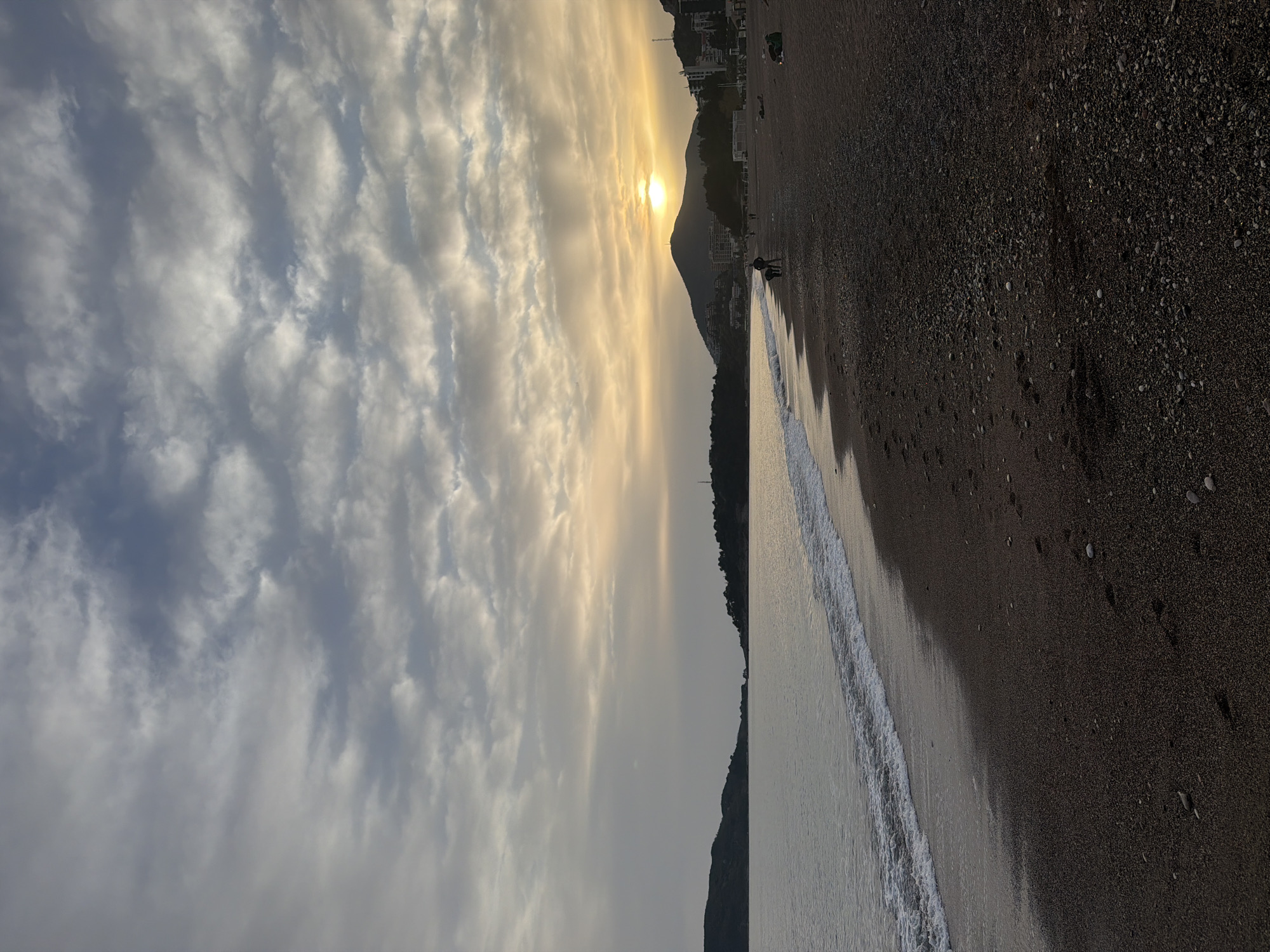 The image shows a beach scene in Rafailovići, Montenegro at sunset. A beach with dark sand and pebbles stretches along the right side, meeting the water's edge where gentle waves roll in. On the horizon, a mountain obscures part of the setting sun, which casts a golden glow across the sky. Altocumulus clouds fill the sky above, reflecting the sunlight. Buildings are visible along the coastline in the distance. Two people walk along the beach near the water.