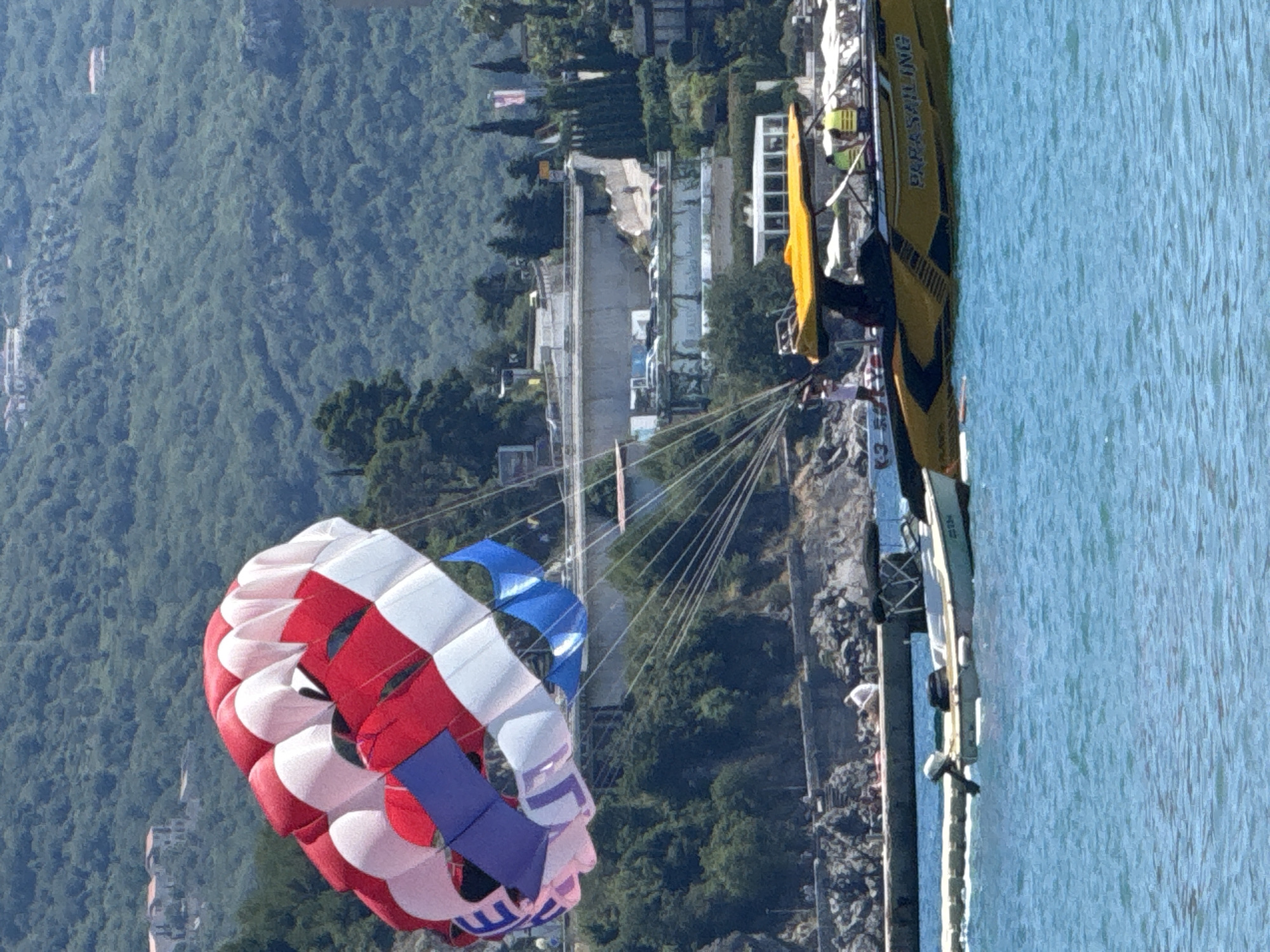 The image shows a parasailing boat with "PARASAILING" written on its side, in the waters off the coast of Budva, Montenegro. The boat is yellow with black and yellow accents. A red, white, and blue parasail is deployed in the air, attached to the boat. A person appears to be standing on the boat. In the background, there is a lush green, tree-covered hillside with buildings and structures on it, and the blue sea in the foreground. A smaller white boat is docked near the shore.