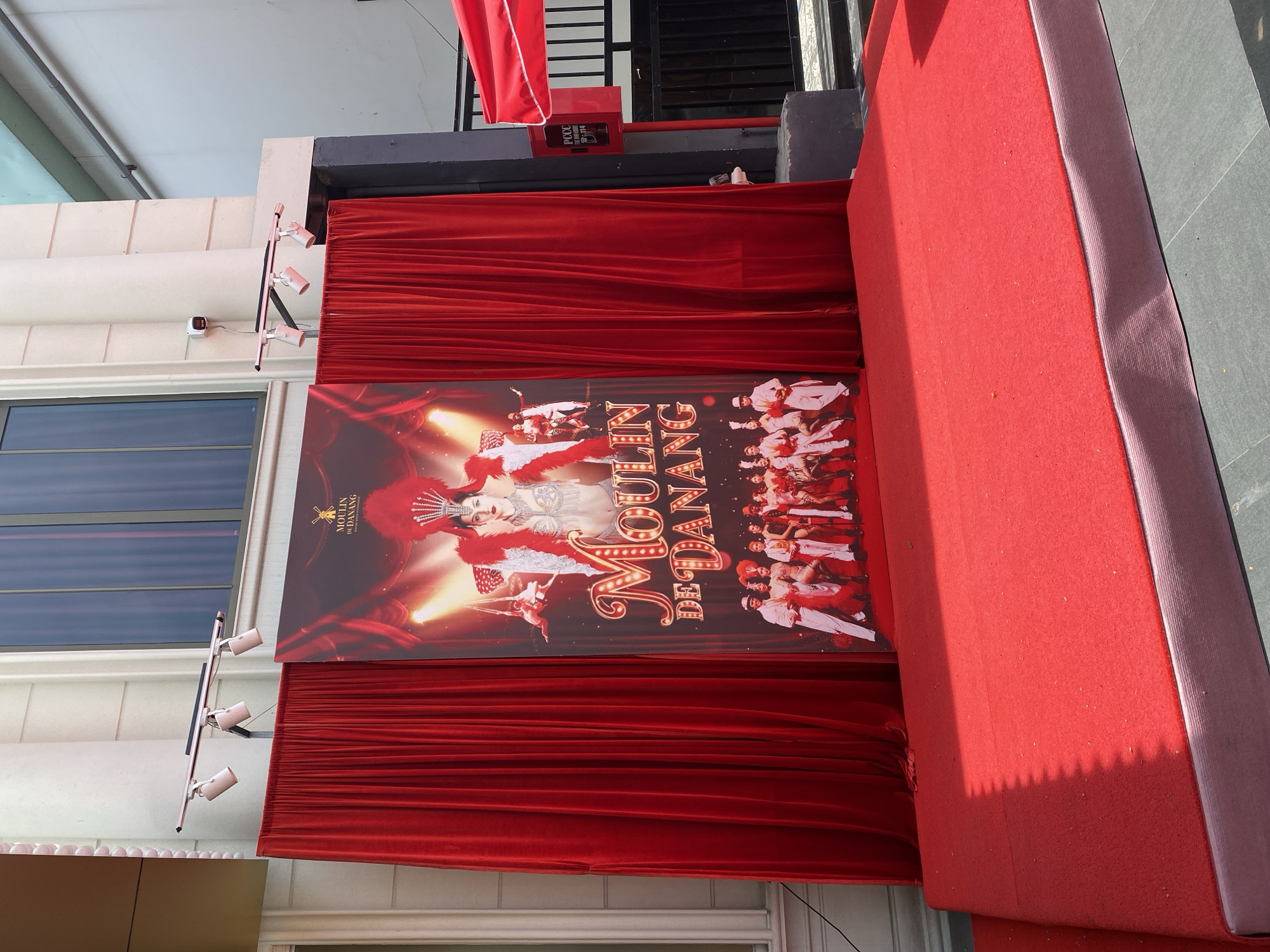 This image shows the entrance to "Moulin De Danang" in Da Nang, Vietnam. A red carpet leads up to a stage adorned with red velvet curtains. Behind the curtains is a large poster featuring a showgirl in a headdress and feathers, with performers in acrobatic poses and a chorus line of dancers at the bottom. The poster is illuminated by spotlights mounted above the curtains. The building itself is light-colored with large windows. To the right of the entrance, a black metal gate is visible, along with a red awning and a red box mounted on a post, possibly a call box or a fire alarm. The overall scene suggests an outdoor entertainment venue or event.