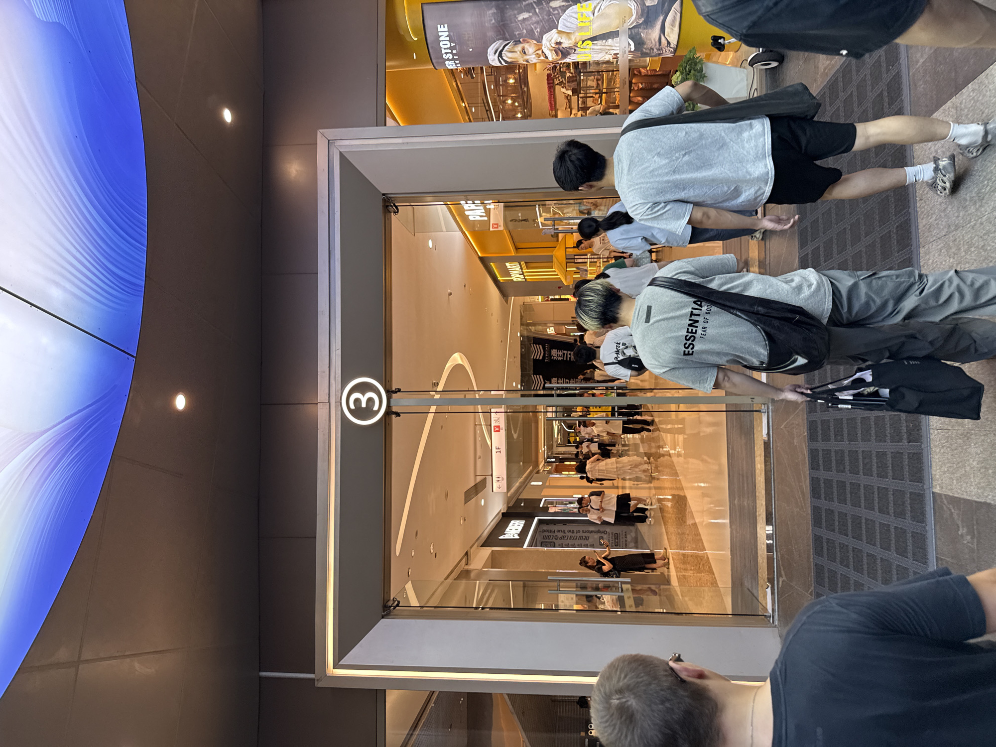 The image shows the entrance to a shopping mall, labeled "3", in Haizhu District, China. Several people are walking into the mall. Inside, various shops and displays are visible, including a "New Era Cap Com" store and a "POPMART". The shops appear well-lit. The exterior features modern architectural elements.