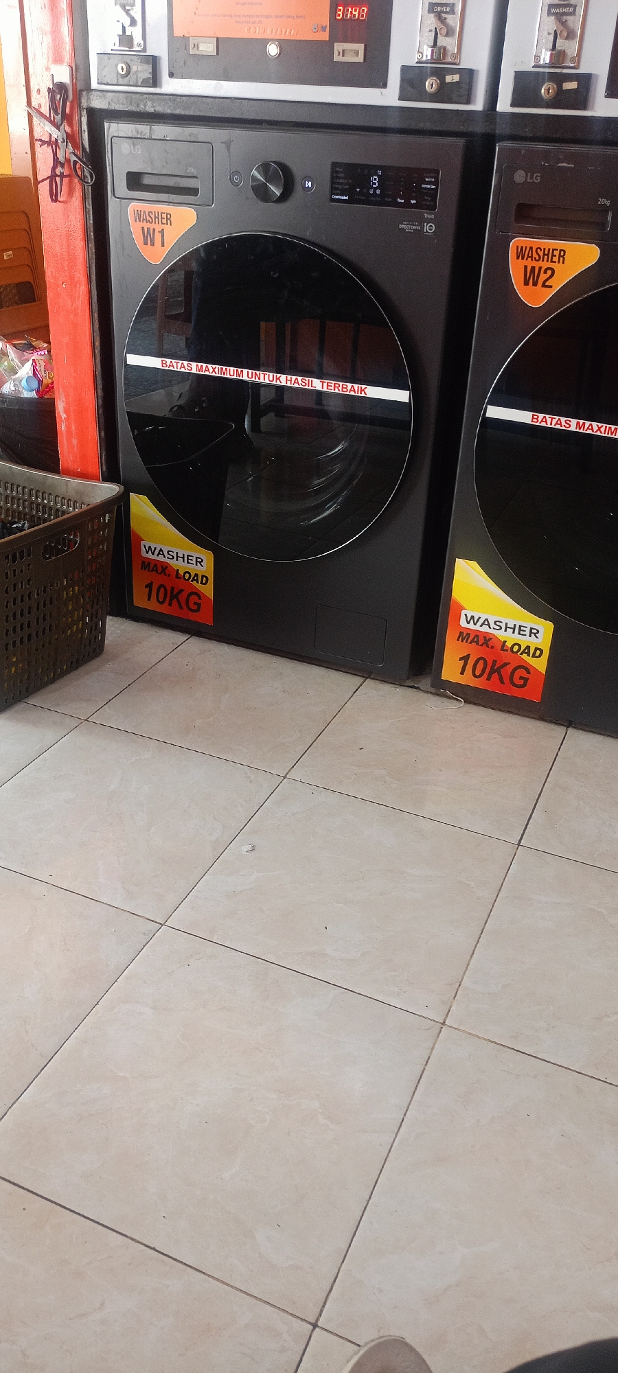 The image shows a close-up of two LG washing machines in a commercial laundry facility, likely located in Mekar Rahayu, Indonesia. The washing machines are dark gray with large glass doors. A sticker on the left washing machine reads "WASHER W1 MAX. LOAD 10KG". A similar sticker on the right machine reads "WASHER W2". A white sticker across the glass door of the left machine states "BATAS MAXIMUM UNTUK HASIL TERBAIK", which translates to "MAXIMUM LIMIT FOR BEST RESULTS". In the foreground, to the left of the washing machines, is a dark brown plastic laundry basket. The floor is tiled with light beige square tiles. In the background, a red-painted wall is visible, with a pair of scissors hanging from it. Above the washing machines, various control panels and slots for coin insertion are visible, consistent with a self-service laundromat.
