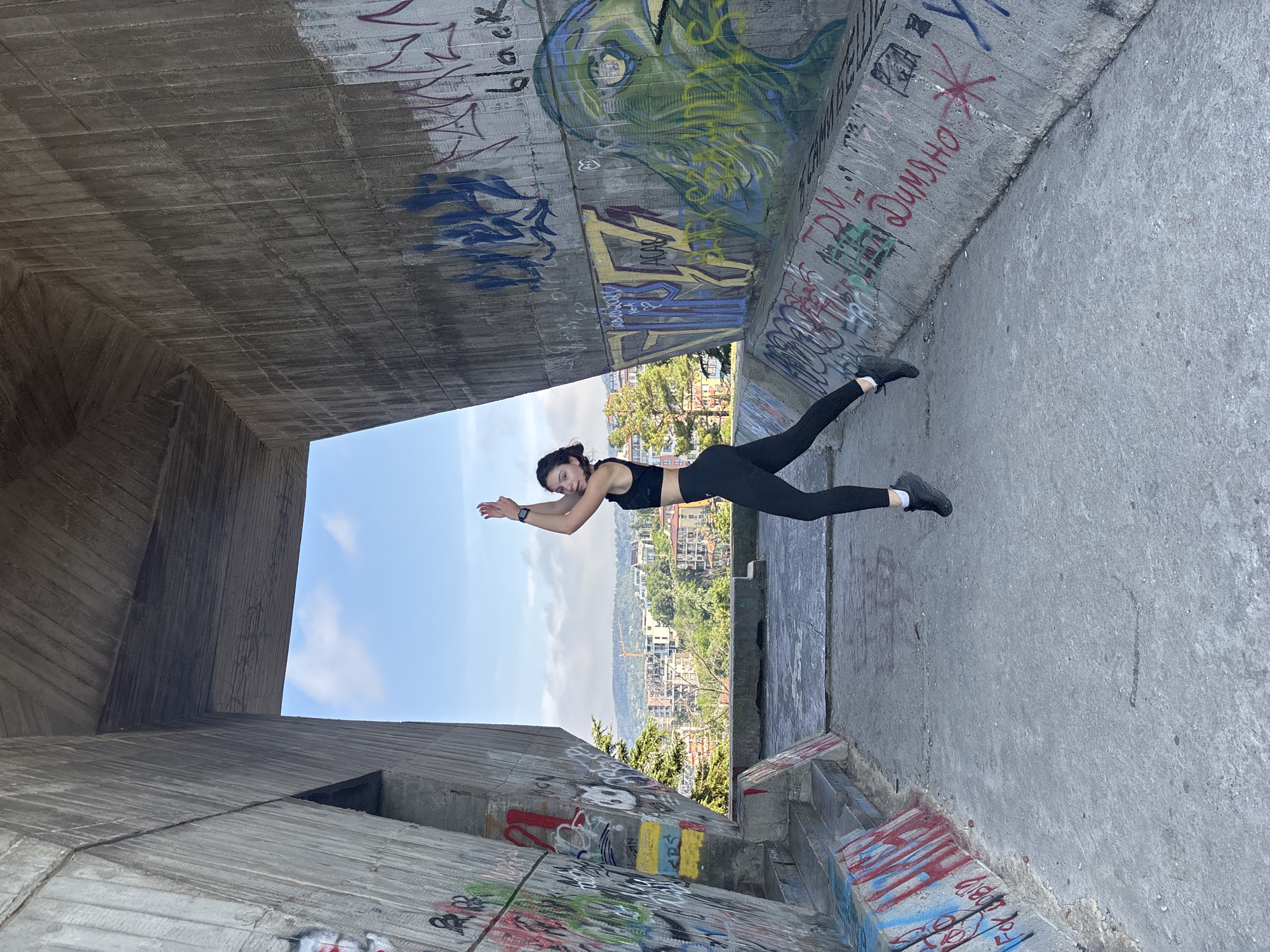A young woman in athletic wear is captured mid-air in a jump within a concrete structure adorned with graffiti. The structure appears to be part of an urban landscape, possibly a bridge or an overpass, with a view of buildings and greenery in the background. The scene is set in the city of Varna, Bulgaria, suggested by the Cyrillic script visible on the graffiti. The woman, with dark hair pulled back, is wearing a black sports bra and leggings, her arms raised above her head in a dynamic pose. The concrete walls are heavily tagged with various colors of spray paint, including abstract designs and text. The ground is concrete, showing signs of wear and tear. The sky is blue with scattered clouds.
