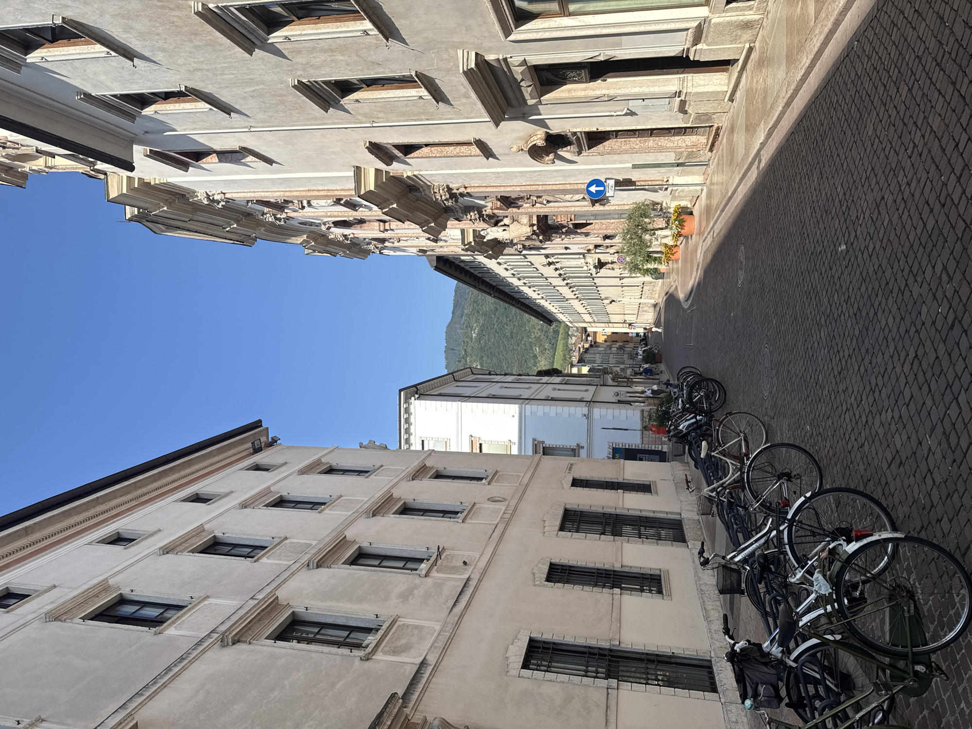 The image depicts a street scene in Trento, Italy. The street is paved with cobblestones and lined with historic buildings on both sides. On the left, a cream-colored building features multiple windows with dark frames and decorative stone surrounds. On the right, a building with an ornate facade, exhibiting classical architectural elements such as columns, pediments, and sculptures, dominates the view. The sky is clear and blue.
Several bicycles are parked along the left side of the street. In the background, the street curves and leads towards a hillside covered in greenery, with more buildings visible along the route. A blue street sign with a white arrow pointing upwards is mounted on a pole near the right side of the street. Potted plants with green foliage and yellow flowers are placed along the sidewalk in front of the ornate building. Two individuals are visible walking in the distance on the right side of the street. The scene appears to be set during the daytime under bright sunlight, casting shadows on the street and buildings.