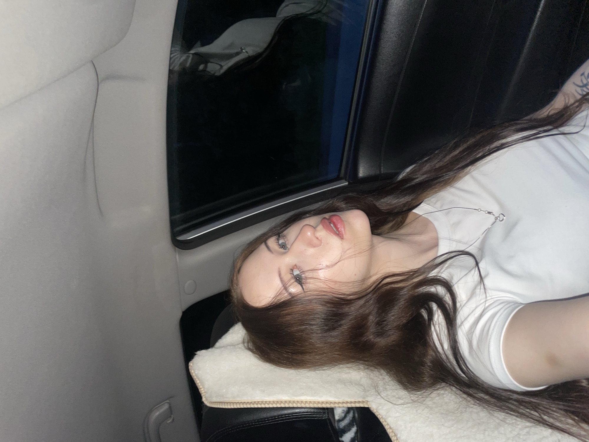 A young woman with long, brown hair is seated in the passenger seat of a car. She is looking towards the camera with light blue eyes and has dark eyeliner. She is wearing a white t-shirt and a silver necklace with a heart-shaped pendant. A fluffy white car seat cover is visible behind her. The car interior is dark, with the window reflecting the night sky and trees outside. The location is not specified within the image or its context.