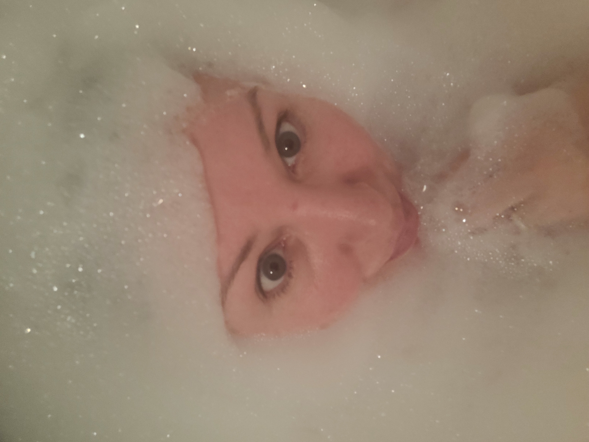 A woman's face is visible amidst a mass of bubbles, likely in a bathtub. Her eyes are open and directed upwards. The background is obscured by the bubbles. The image provides no data to determine if the photo was taken in Izhevsk, Russia.