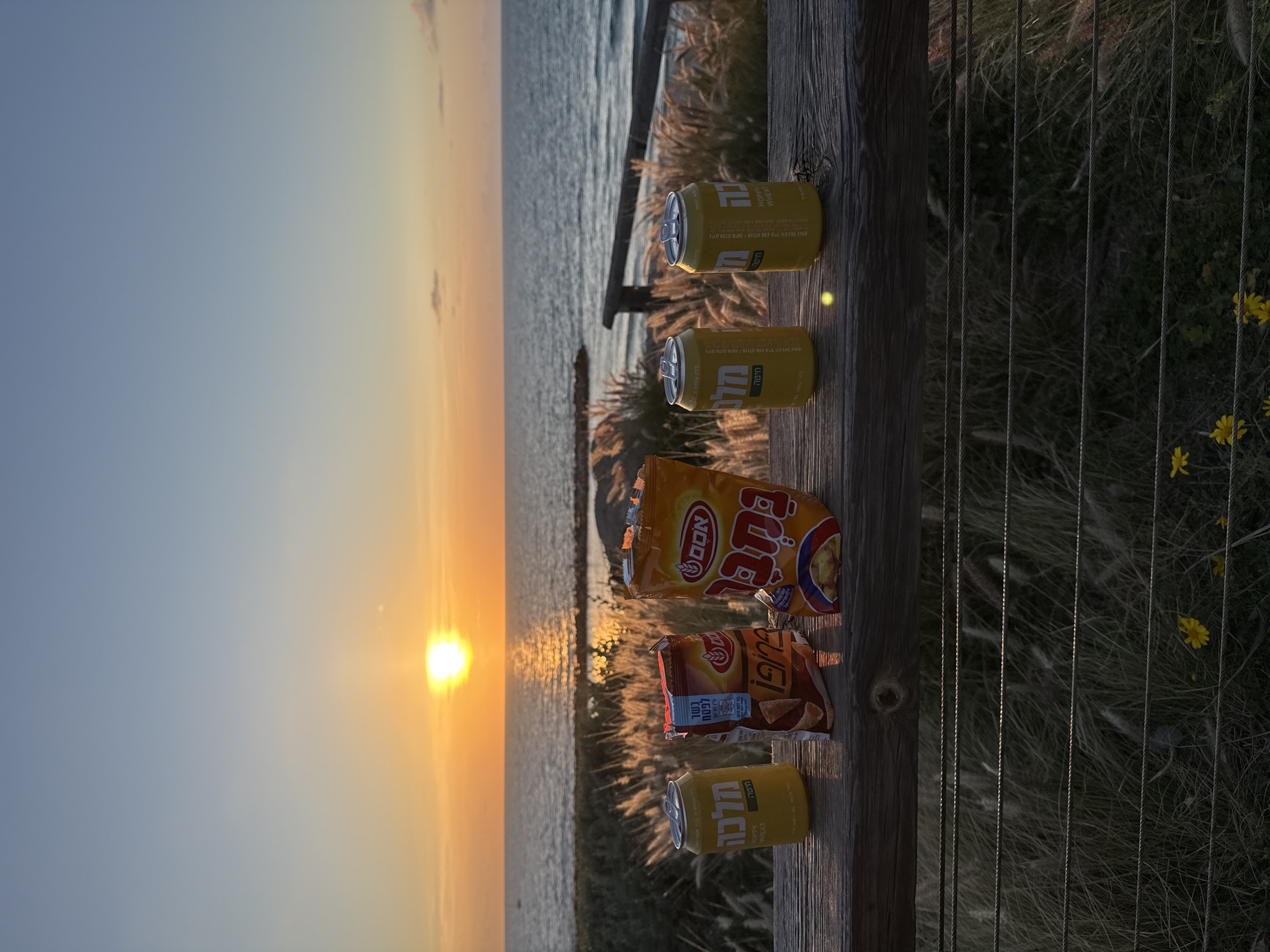 The image shows a scenic overlook at sunset, likely in Tel Aviv, Israel, given the Hebrew writing on the snack and beer containers. A wood railing is in the foreground holding three cans of "Malka" beer and two bags of chips. The backdrop features the Mediterranean sea, reflecting the bright sunset. The surrounding area appears to be vegetated with tall grass and some yellow flowers.