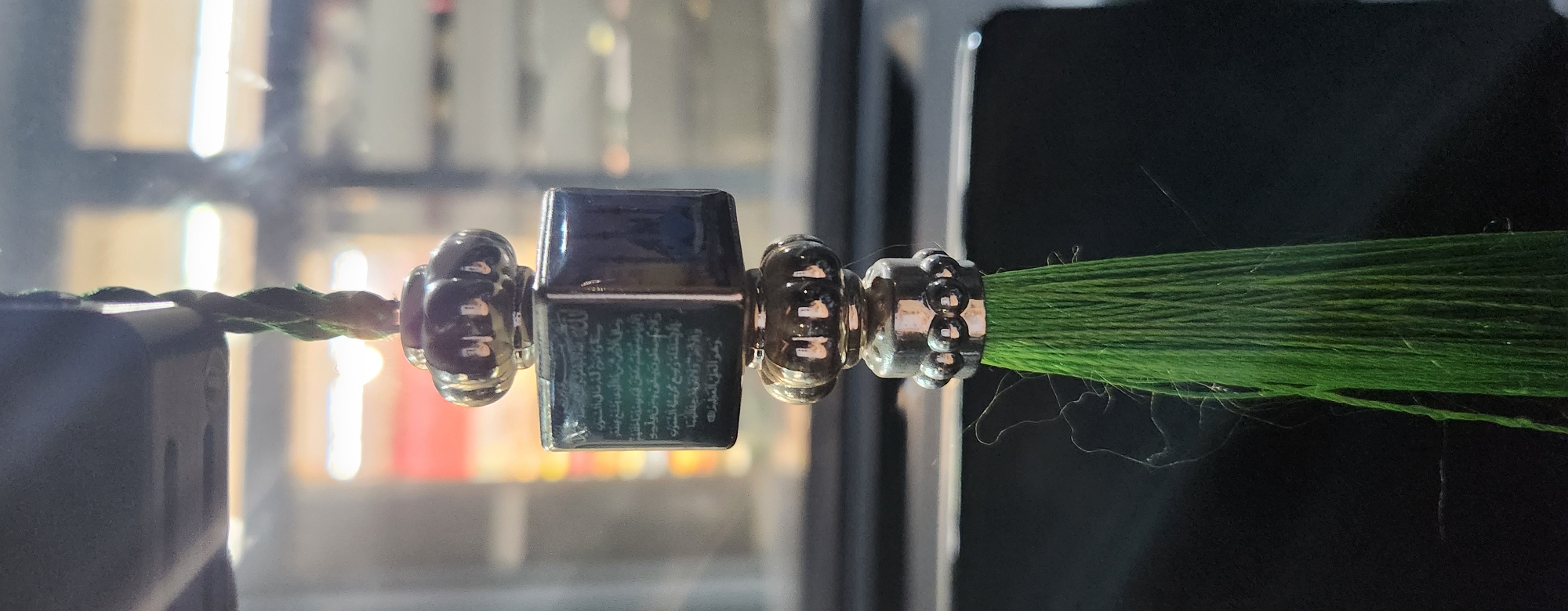 A close-up, vertical shot shows a green tassel hanging in front of a blurred background that appears to be a window with distant lights and reflections. The tassel is attached to a dark, square object with Arabic calligraphy etched onto its visible face. Above this square object is a decorative, metallic bead with a scalloped design, and below it are two more metallic beads, the lower one being larger and more detailed. The green tassel itself is composed of many thin strands. The scene is likely an interior space, possibly a car or a room, with the focus solely on the hanging decorative item. There are no people or animals present in the image. The specific location in Kazan, Russia, cannot be determined from the provided image alone.