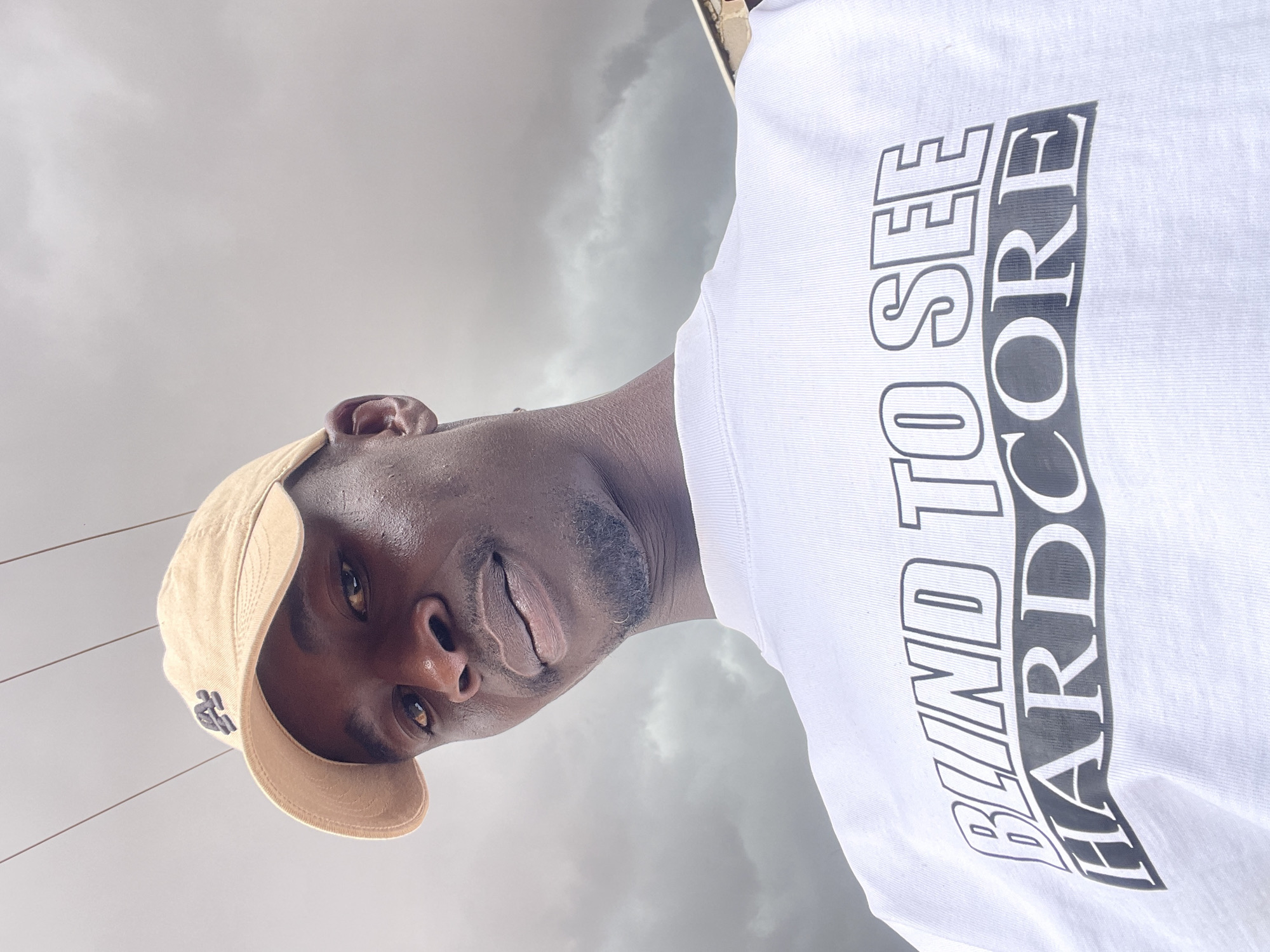 A man is pictured from a low angle against a cloudy sky. He is wearing a tan baseball cap and a white t-shirt with the words "BLIND TO SEE HARDCORE" printed in a shadowed font. The man has dark skin, dark eyes, a short beard, and appears to be looking directly into the camera. Thin wires are visible in the top-left of the frame. The location is unknown, but the prompt states it is in the city of Jos, Nigeria.