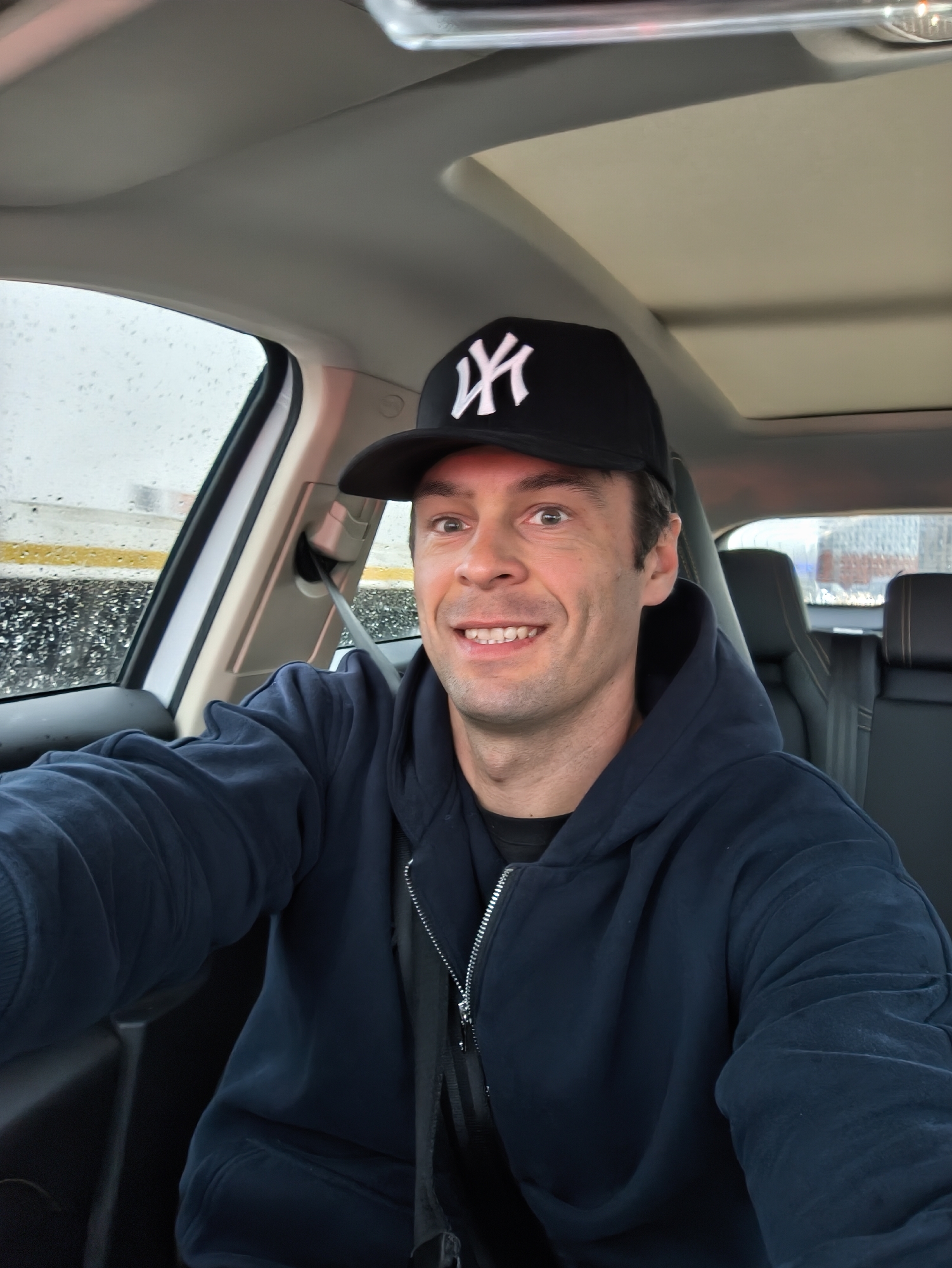 A man wearing a black New York Yankees baseball cap and a navy blue zip-up hoodie is taking a selfie while sitting in the driver's seat of a car. He is wearing a seatbelt. Rain streaks the car windows, obscuring the view outside. Through the front window, there are indistinct objects and yellow markings which suggests an urban environment. Through the rear window, indistinct buildings can be seen. Given the reference to the city of Moscow, Russia, the location can be inferred to be there, but there are no details in the photo to confirm this.