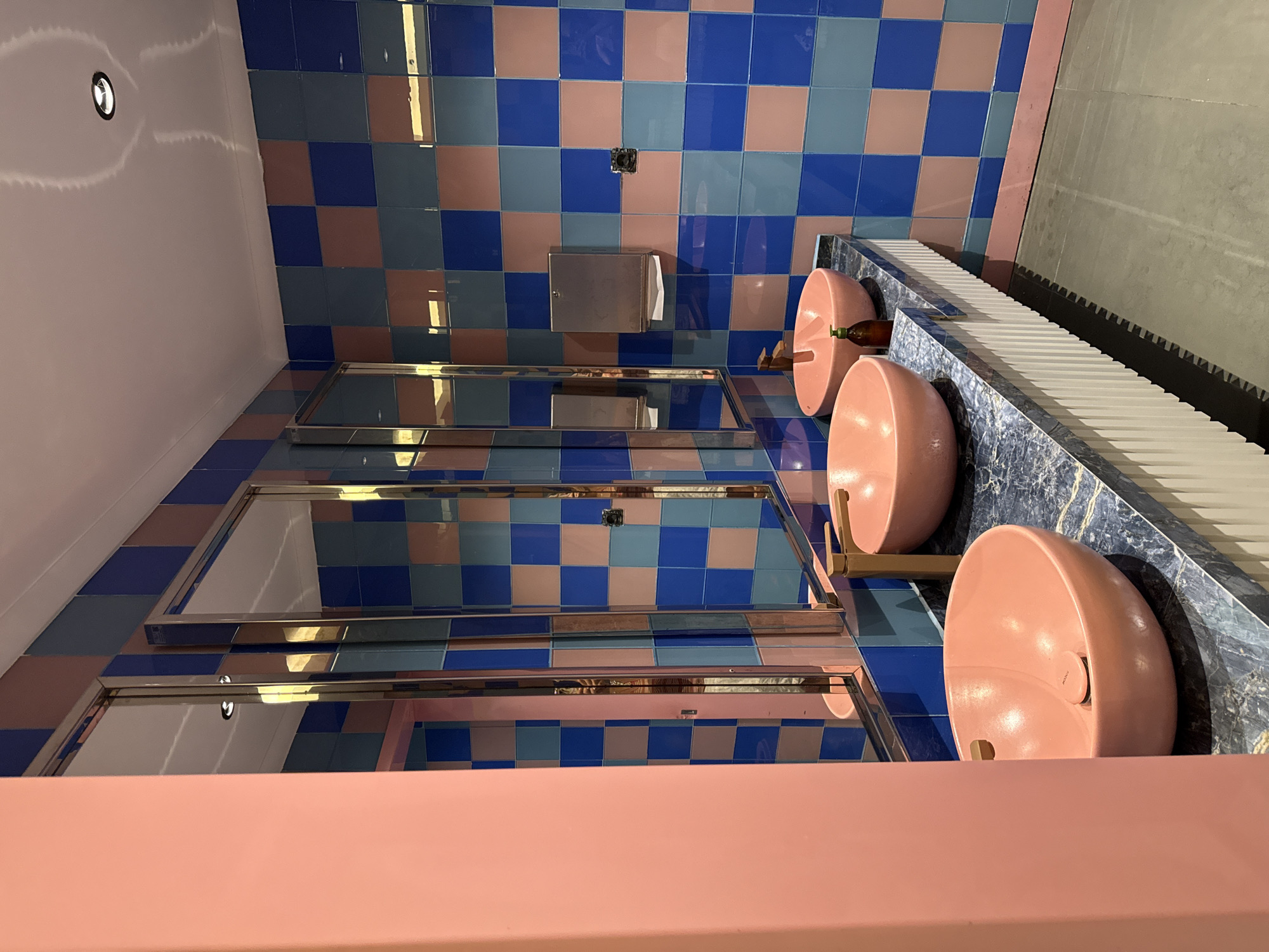 This is a forensic, matter-of-fact description of a modern public restroom, possibly located in Bodrum, Turkey, with a vibrant and distinct design aesthetic. The walls are tiled in a checkered pattern of bright blue and a muted pink. A large, salmon-pink pillar is visible on the left, adding to the colorful scheme.
The central feature is a long vanity countertop with a dark, marbled pattern. Mounted on the countertop are two large, salmon-pink vessel sinks, each equipped with a brushed gold faucet. Between the sinks, a small brown glass bottle, likely containing soap or lotion, is placed.
Above the vanity, a series of large mirrors are set into chrome frames, reflecting the checkered walls and ceiling. A metallic paper towel dispenser is mounted on the wall behind the sinks.
The floor is a neutral gray, and the ceiling is white with a single recessed light fixture visible. The overall impression is one of a contemporary and stylish, if somewhat unconventional, restroom design. No people are present in the image.