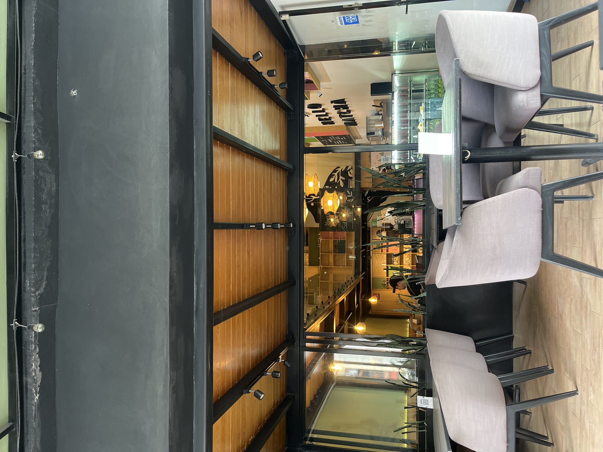 The image shows an interior view of a business, potentially a café or restaurant, in San Bernardino Tlaxcalancingo, Mexico. The camera perspective is from the entrance, looking inside. The entrance structure features a dark gray metal frame with wooden beams, which have small track lights attached. Inside, there is a partially visible counter with the words "HELLO MATCHA" displayed on it. The interior design includes shelving with various items, suggesting the space is a retail or dining establishment. The lighting is warm, and there are some visible plants decorating the interior. In the foreground, there are several chairs with light gray upholstery positioned around dark-colored tables, set up for customers. One person is visible inside, near the back of the business. A glass partition or window allows a view into the space.