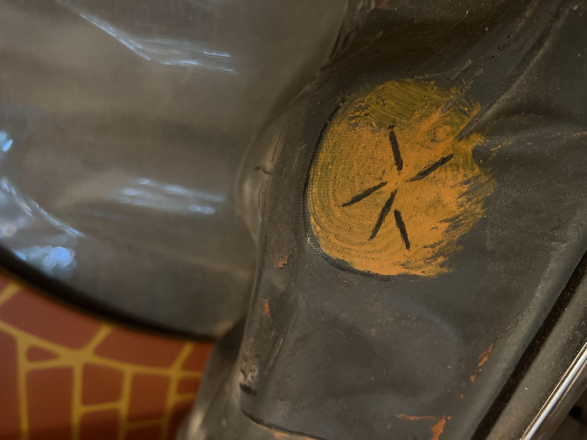 A close-up, angled shot focuses on a portion of a dark, possibly ceramic or painted object. Dominating the center of the frame is a bright orange circular marking with thick black lines radiating outwards, resembling a stylized sun or star. The orange paint is roughly applied, with visible brushstrokes and some areas of lighter and darker pigment. The surface surrounding the orange marking is primarily dark gray or black and appears somewhat textured and worn, with small flecks of reddish-brown paint visible in places, suggesting age or damage. In the upper left of the image, a section of a red and yellow patterned surface is visible, resembling bricks or tiles, set against a curved, reflective gray surface. The lighting creates highlights and shadows, particularly on the curved gray area, hinting at a glossy or smooth texture. The overall impression is of a detail from a larger, possibly decorative or toy-like object.