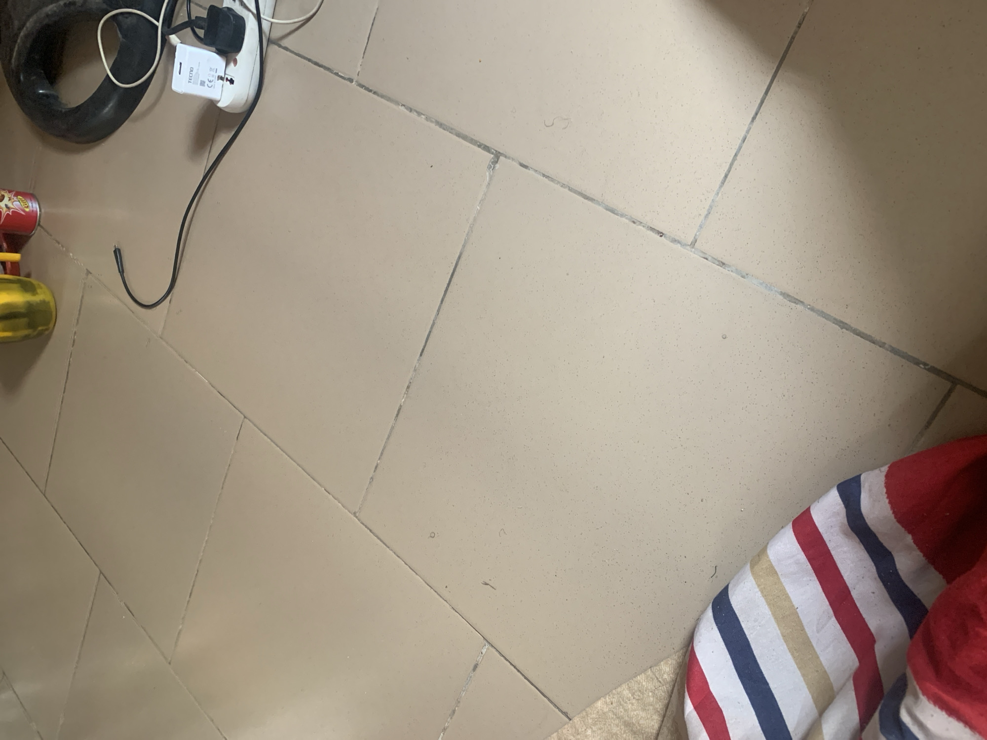 The image depicts a tiled floor, appearing to be in a residential or commercial space, likely within Jauro Yinu, Nigeria. The tiles are a neutral, sandy beige color with visible grout lines. In the upper right quadrant of the image, a white electrical adapter is plugged into a power strip, which is connected to a black cable. A black electrical plug is inserted into the adapter. To the left of the adapter, a black cylindrical object, possibly a tire or a large rubber ring, is present. Further to the left, a portion of a yellow cylindrical object and a red can are visible. In the lower left corner, a fragment of a striped fabric, featuring red, white, blue, and beige stripes, is visible. The overall scene suggests a common household or office environment.