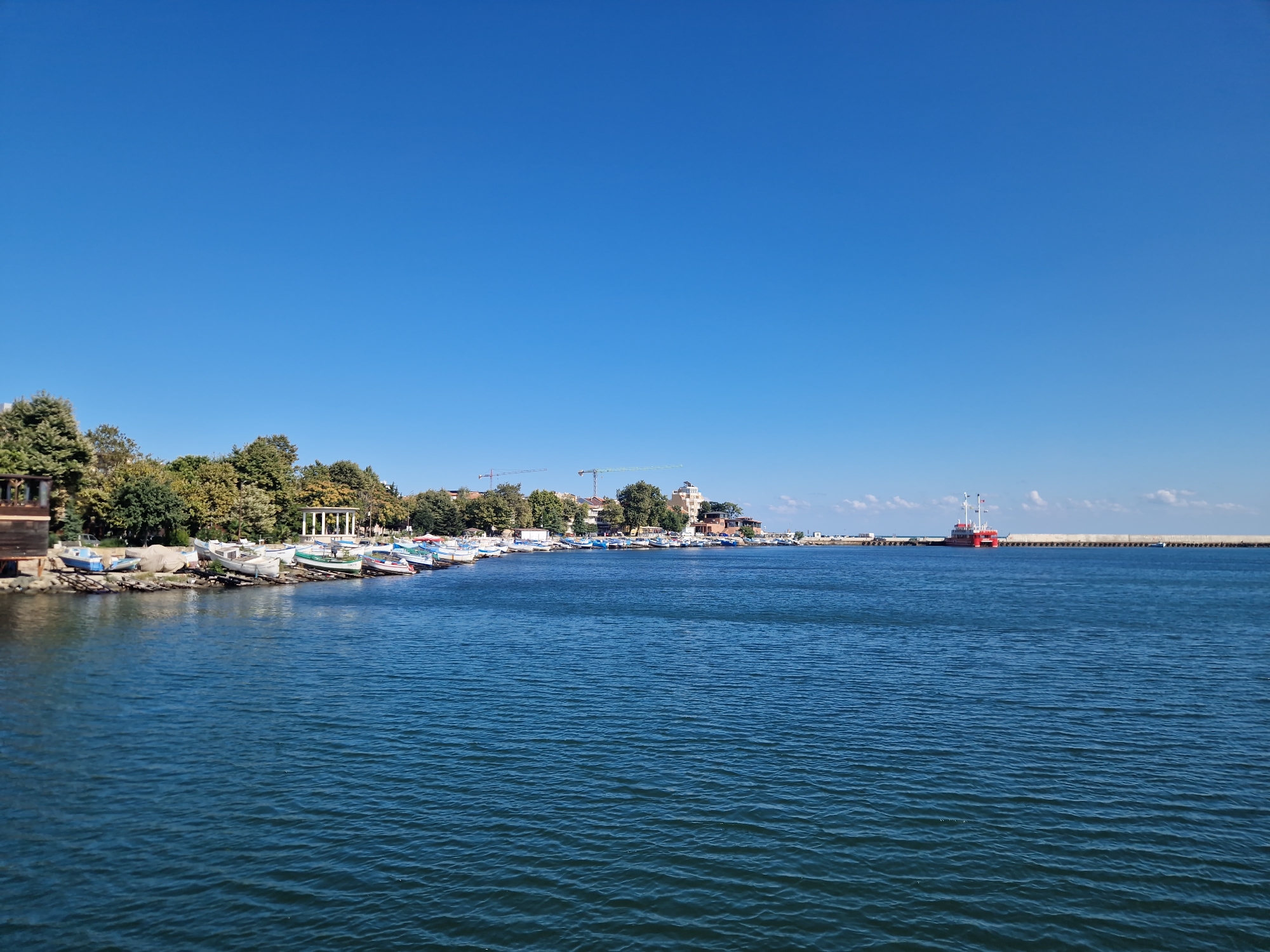 The image shows a scenic view of the coastline in Pomorie, Bulgaria, on a clear, sunny day with a bright blue sky. The foreground is dominated by the deep blue sea, with gentle ripples on its surface. Along the shoreline, a line of small fishing boats are docked, many of them white, resting on the shore or partially in the water. Behind the boats, a lush green line of trees and vegetation forms the backdrop of the land. Several buildings, including what appears to be a small wooden structure on the left and a white, colonnaded building further along the shore, are visible nestled amongst the trees. In the distance, the silhouette of a port or harbor can be seen, with a breakwater stretching into the sea. A prominent red and white vessel is docked at the end of this structure, and construction cranes are visible in the town. The overall impression is one of a tranquil coastal town with active maritime life.