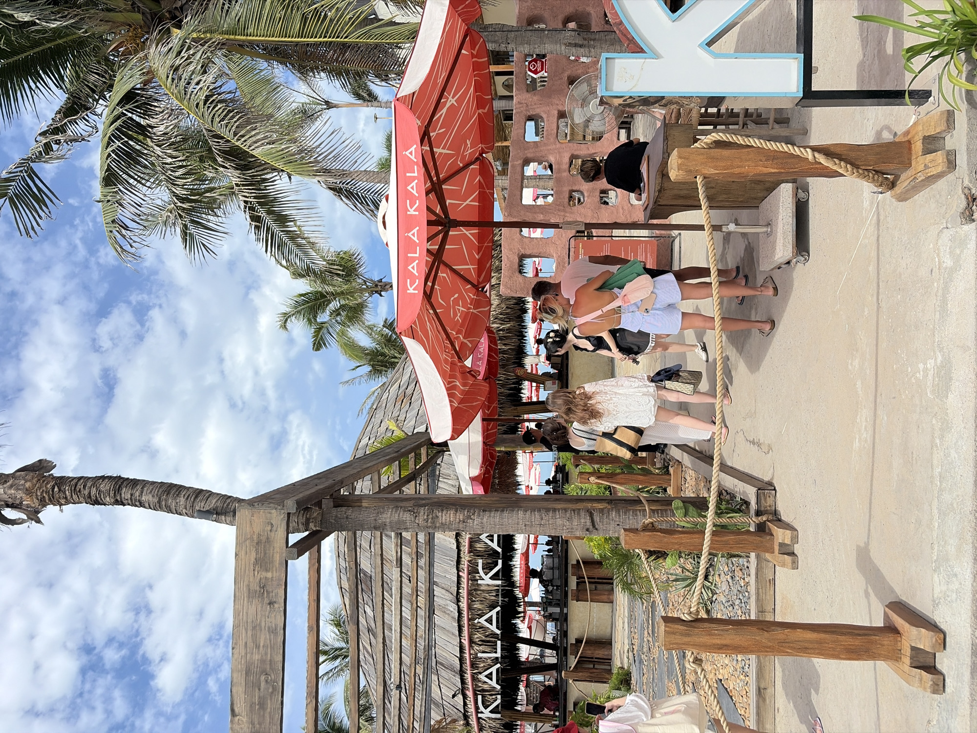 The image shows the entrance to a beachside establishment in Da Nang, Vietnam, with palm trees, a thatched roof building, and umbrellas. Several people are seen walking towards the establishment, some carrying bags. The ground is paved, and wooden posts with ropes demarcate a path. The sky is blue with scattered clouds. A sign with the text "KALA KALA" is visible on a red and white umbrella. The overall scene suggests a casual, tropical atmosphere.