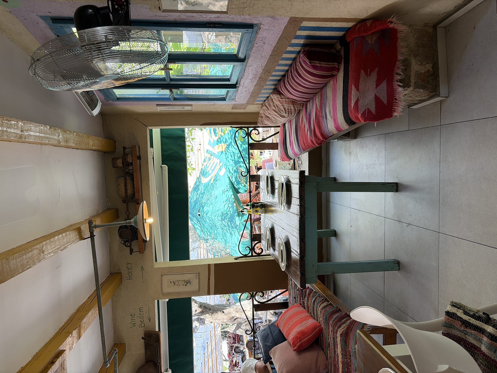 The image depicts the interior of a restaurant in Stari Bar, Montenegro, with a rustic and bohemian ambiance. A long, turquoise wooden table is set with plates and cutlery, suggesting a dining setting. Surrounding the table are two wooden benches upholstered with colorful, patterned textiles and cushions, creating a cozy seating area. The walls are adorned with a mix of textures and colors, and a large fan is mounted on the wall near a blue-framed window. Above the seating area, exposed wooden beams cross the ceiling, and a vintage-style lamp hangs down. Outside the window, glimpses of trees and umbrellas suggest an outdoor patio or garden area. The overall impression is of a charming, relaxed eatery in a historical setting.