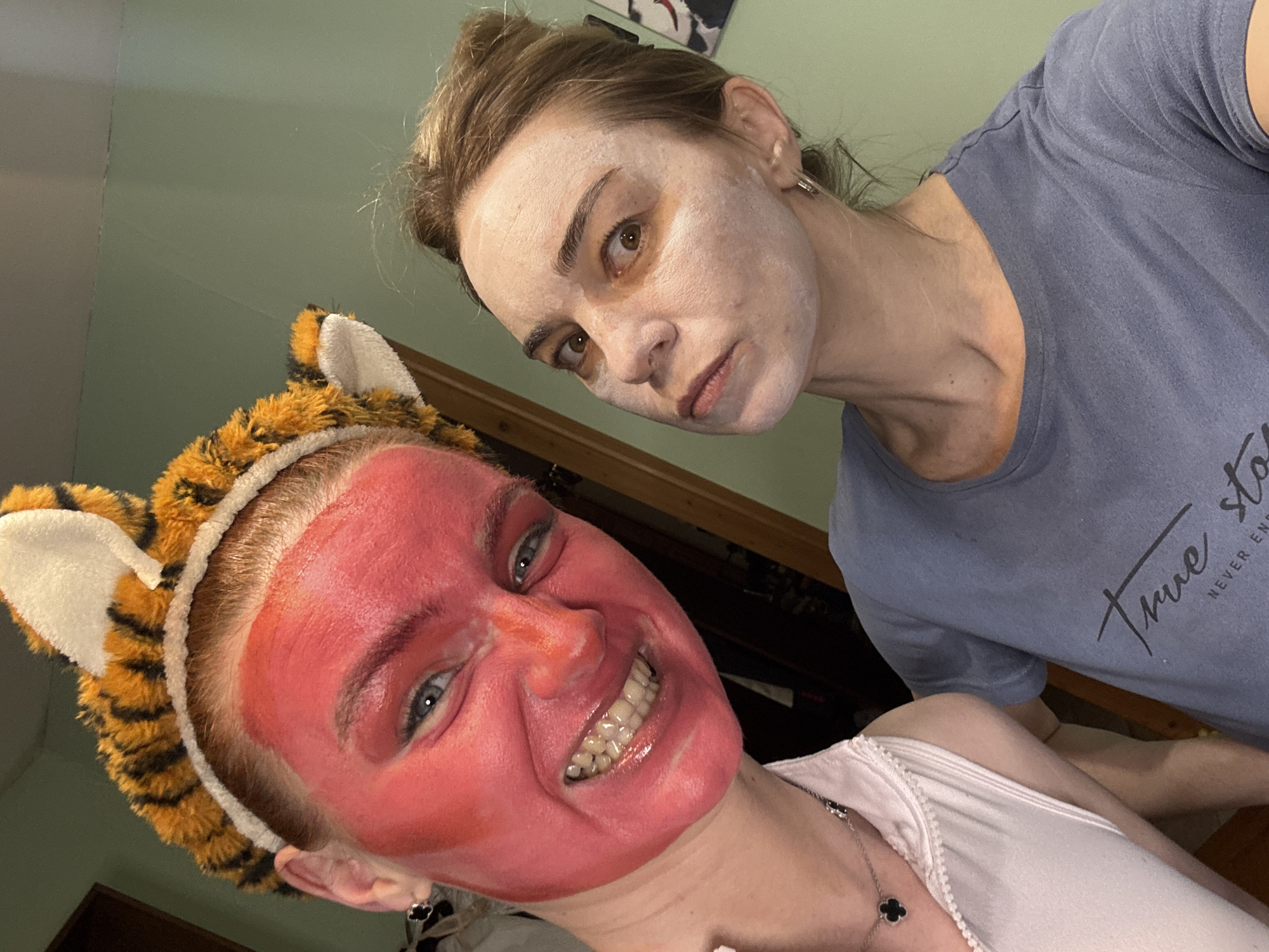 The image depicts two people in an indoor setting. The person in the foreground is wearing a red face mask and a tiger-ear headband, smiling with visible teeth. The person in the background has a white face mask and is wearing a blue t-shirt. The shirt has a "True Story" logo on it. The background is a green wall with a door. The location is a city of Unknown, Unknown.