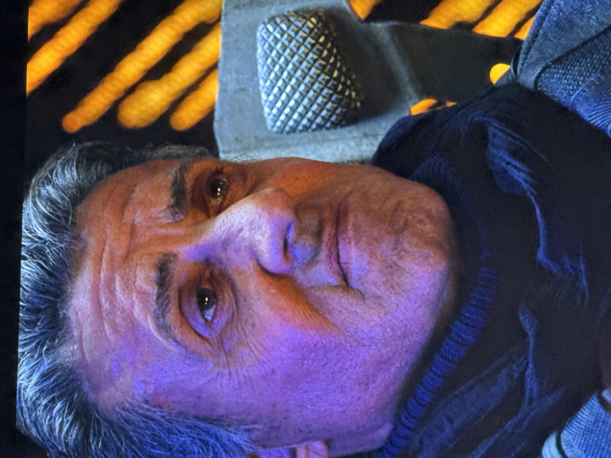 The image is a close-up, head and shoulders shot of a man, likely Sylvester Stallone, with graying hair and wrinkles on his face. He is wearing a dark scarf and a blue plaid jacket. The lighting on his face is predominantly purple and orange, suggesting artificial lighting. In the background, there are blurred out orange and yellow lights, possibly from a city or a building. There is no indication of this image being taken in Riga, Latvia.